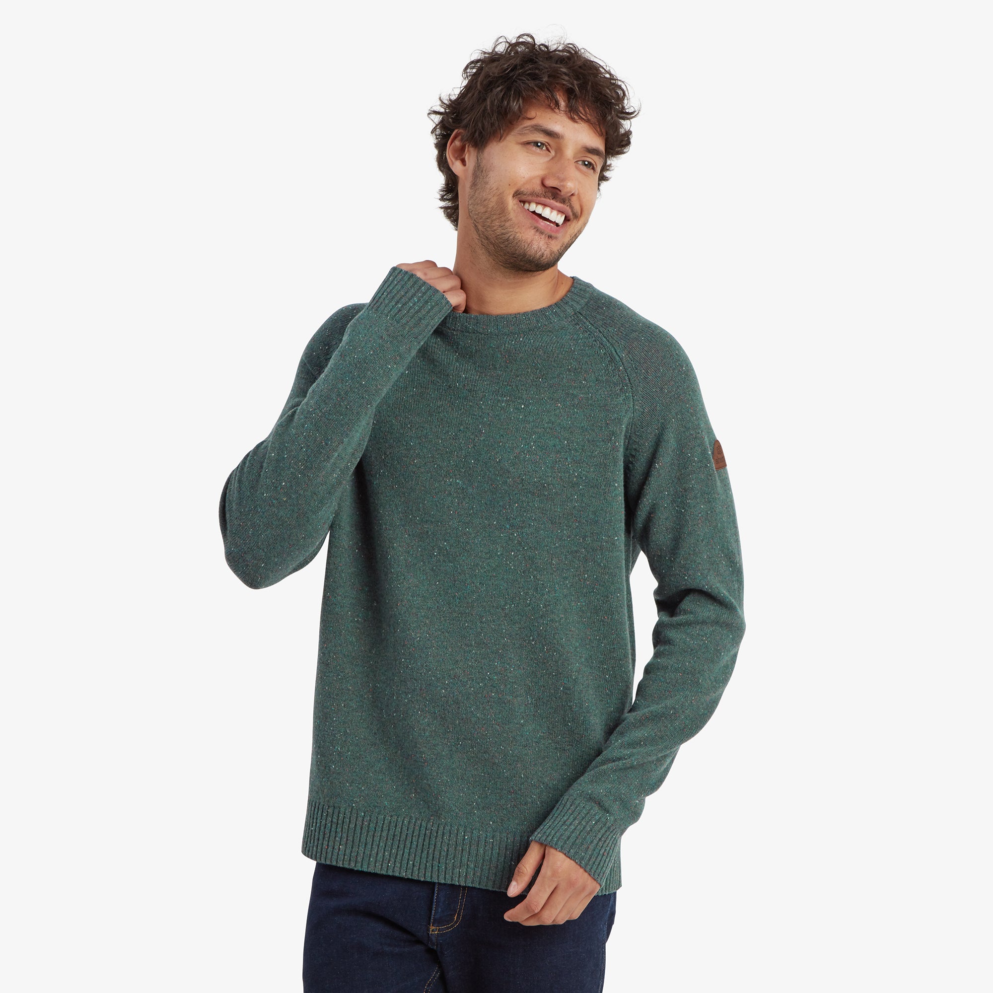 A man is modeling a Sherpa Adventure Gear Kangtega Crew Sweater in Green with subtle speckled details. The sweater fits snugly and has a ribbed hem and cuffs. A brown Sherpa Adventure Gear logo patch is visible on the left sleeve. He is smiling and has one hand near his neck, wearing blue jeans.