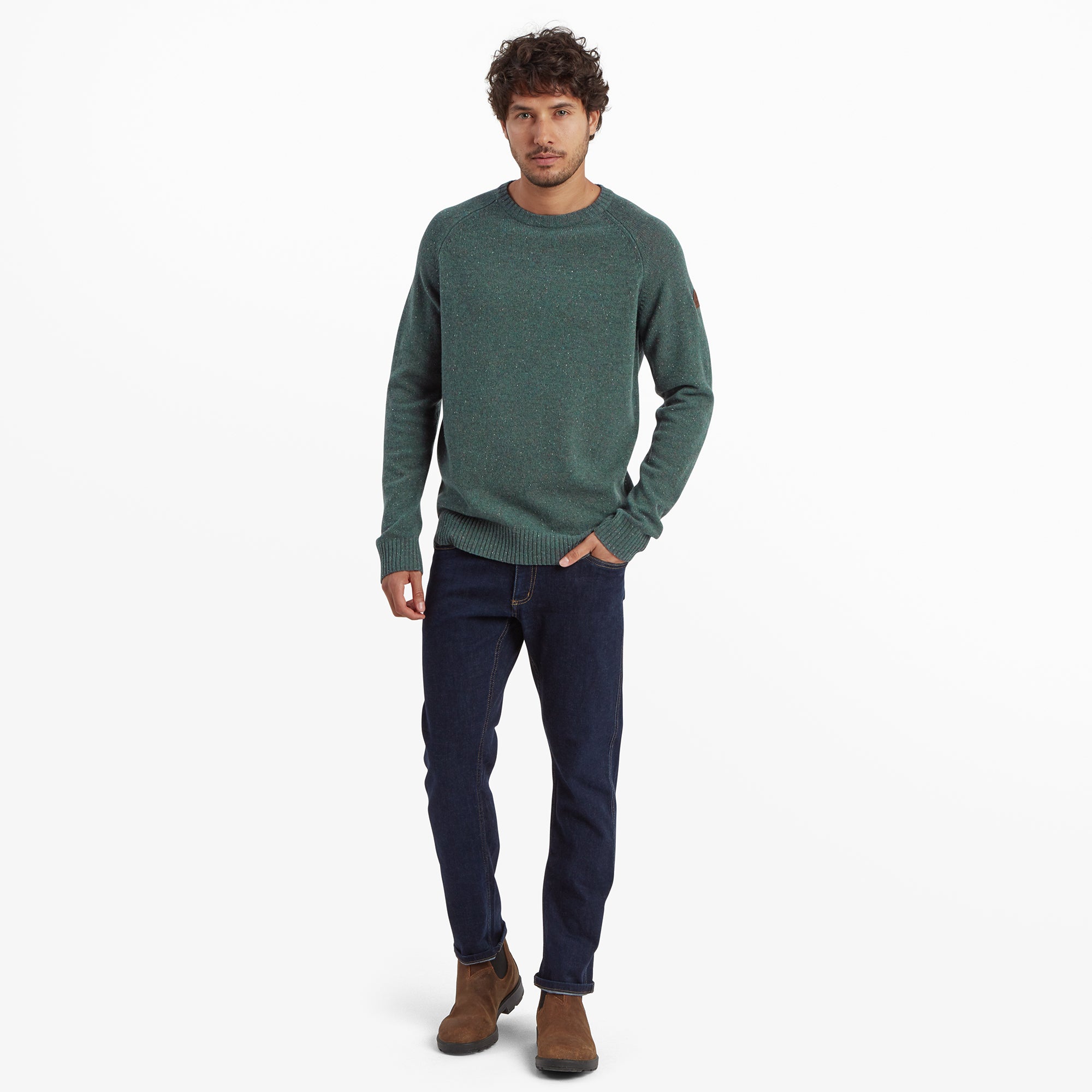 The man is standing upright, showcasing the full outfit. He is wearing the Sherpa Adventure Gear Kangtega Crew Sweater in Green paired with dark blue jeans and brown suede ankle boots. His hands are in his pockets, and the outfit emphasises a casual, outdoorsy style.