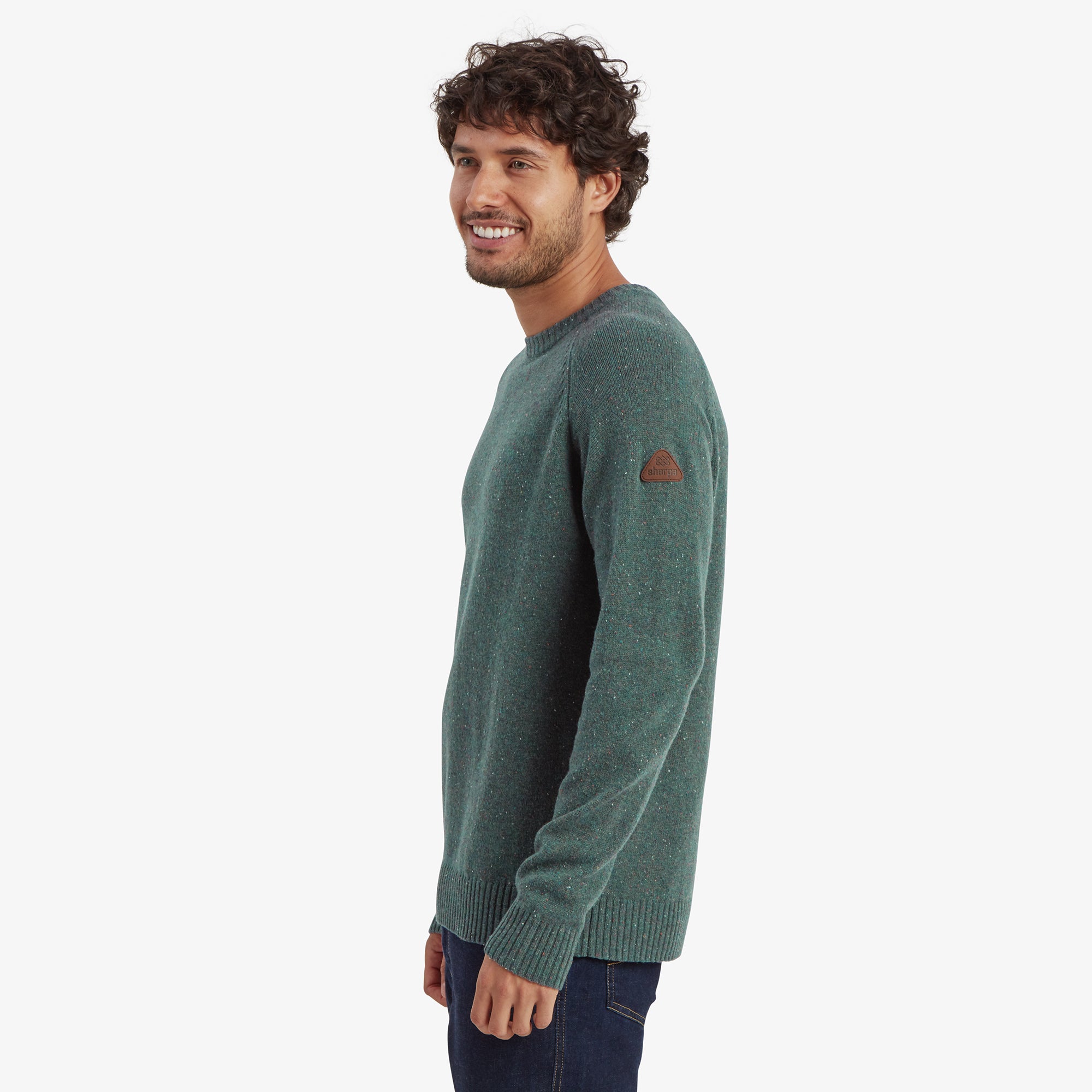 A side view of the man wearing the Sherpa Adventure Gear Kangtega Crew Sweater in Green. The fit is slightly relaxed around the torso. The Sherpa logo patch on the left sleeve is clearly visible. The model is smiling, with his arms hanging naturally.