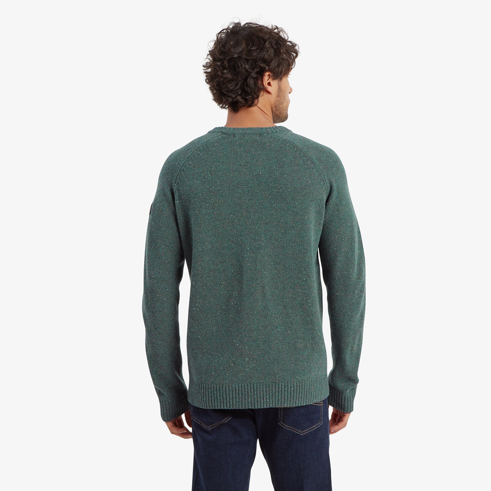 A back view of the Sherpa Adventure Gear Kangtega Crew Sweater in Green, highlighting the clean, uninterrupted design and ribbed detailing at the hem and cuffs. The fit shows slightly dropped shoulder seams. The man is wearing dark jeans, standing with a relaxed posture.