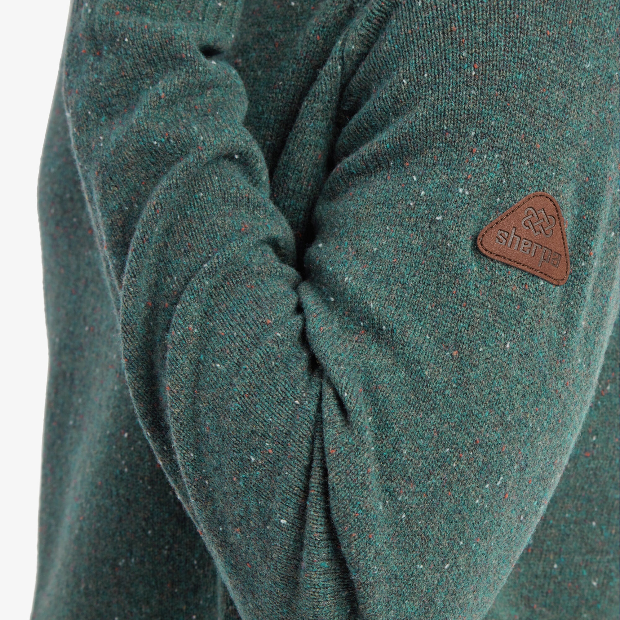 A close-up of the left sleeve of the Sherpa Adventure Gear Kangtega Crew Sweater in Green, focusing on the Sherpa Adventure Gear brown leather patch. The fine speckled texture of the knit fabric is visible, showing red, white, and blue flecks throughout the green base. The stitching of the logo patch and ribbed sleeve cuff is in focus.