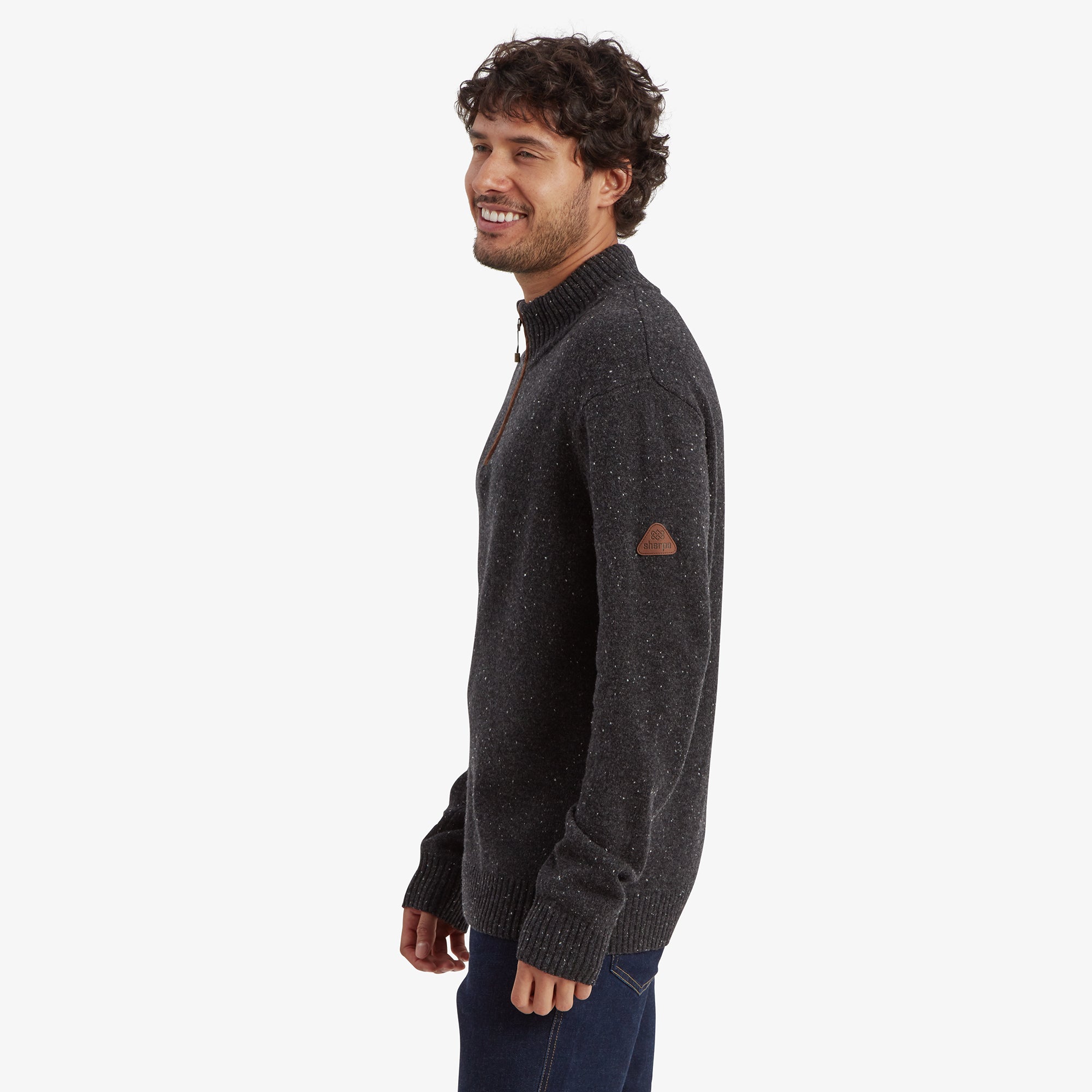 A side view of the man wearing the Sherpa Adventure Gear Kangtega Quarter Zip Sweater in Black. He is smiling and looking over his shoulder, highlighting the sweater's shoulder seams and a small leather Sherpa logo patch on the left sleeve.