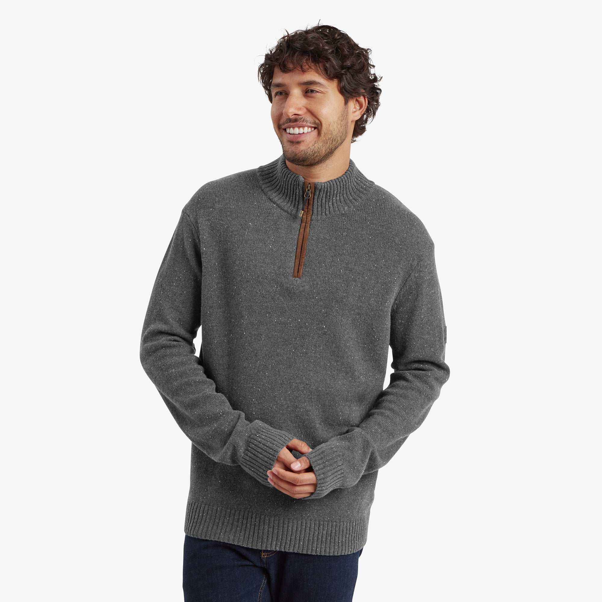 A man wearing a Sherpa Adventure Gear Kangtega Quarter Zip Sweater in Grey with a ribbed high collar. The sweater features subtle speckled detailing and a tan leather zipper pull. He is smiling and holding his hands together, paired with dark blue jeans.