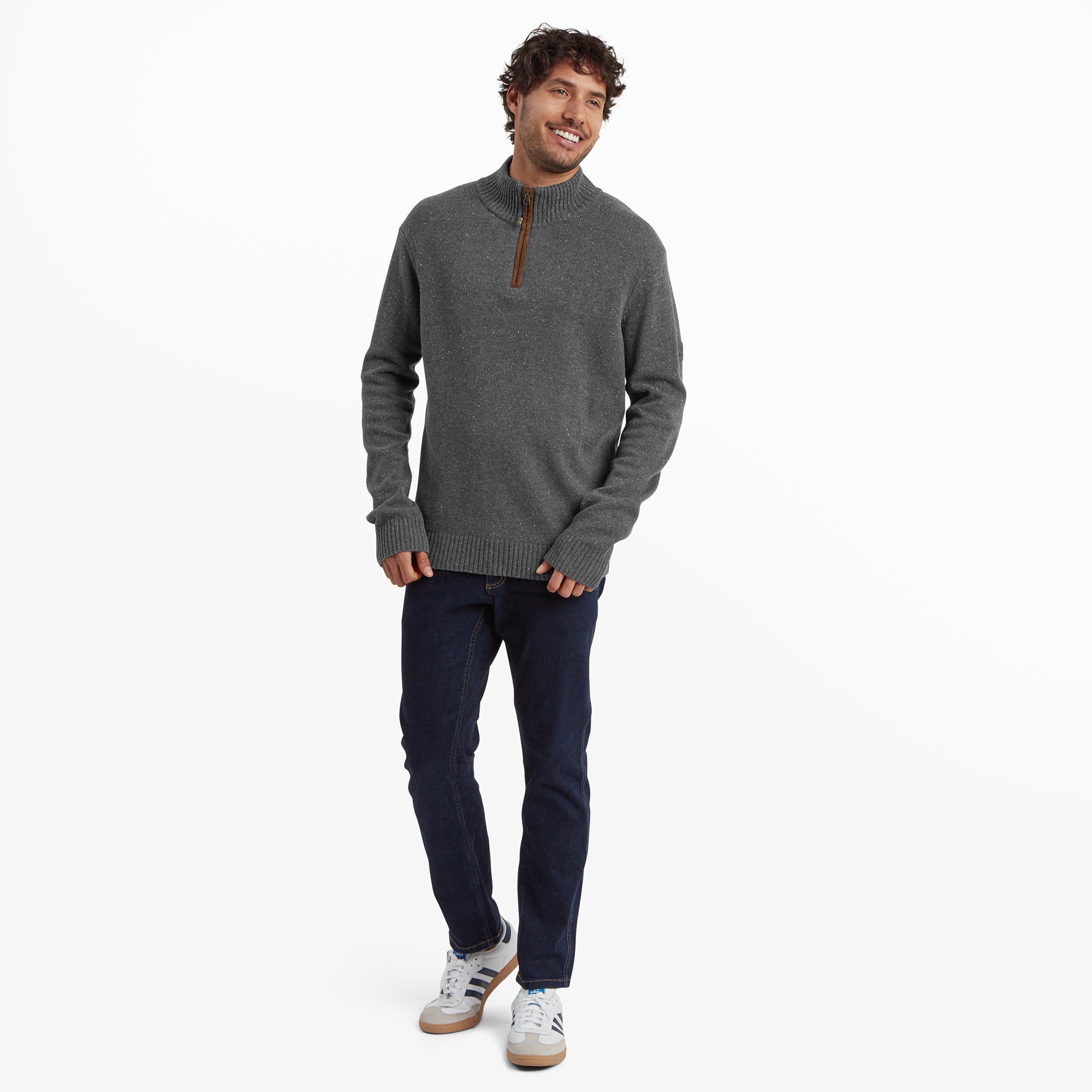 A full-body view of the same man in the Sherpa Adventure Gear Kangtega Quarter Zip Sweater in Grey, standing in a relaxed pose. He pairs the sweater with blue jeans and white trainers, smiling as he tilts his head slightly.