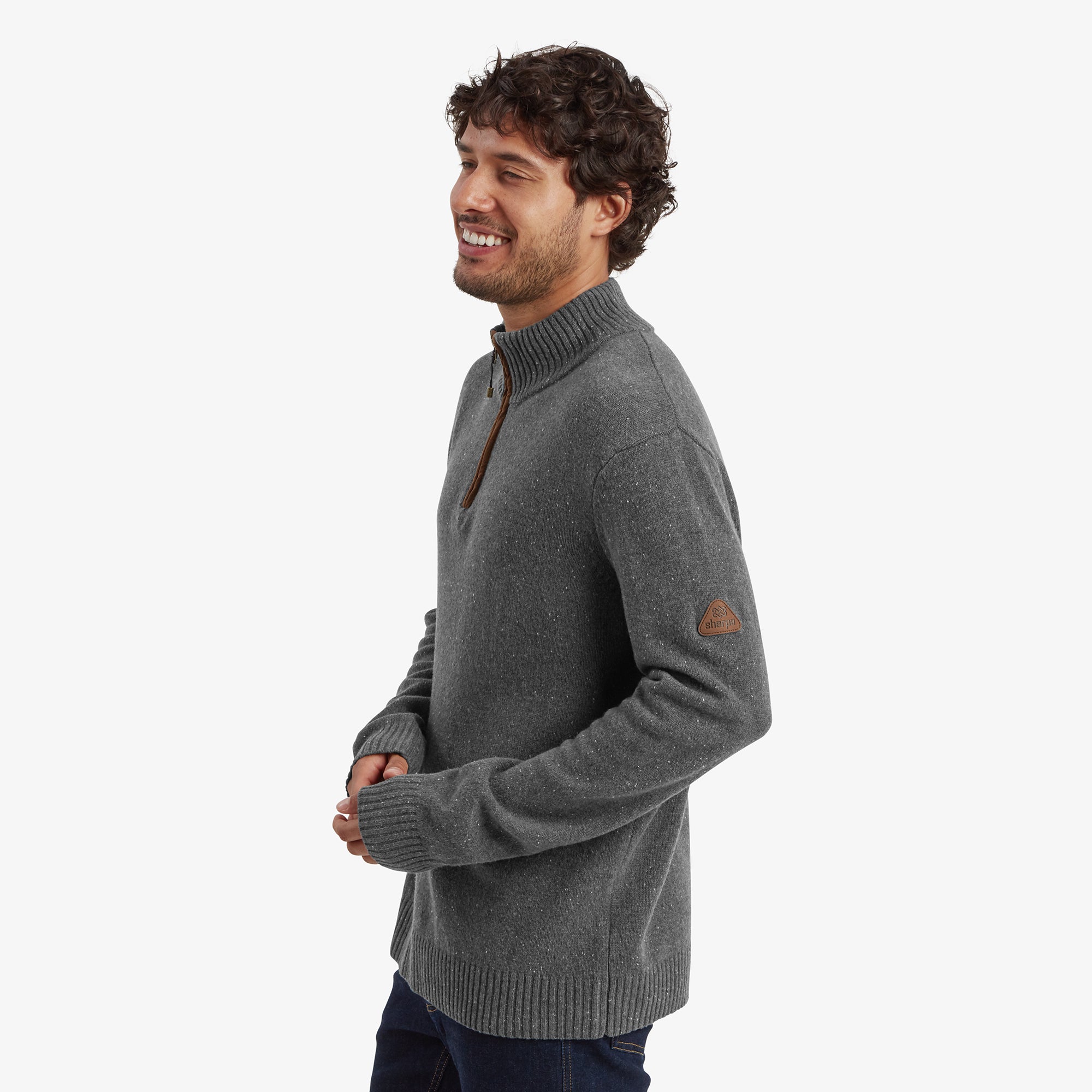 A side view of the man wearing the Sherpa Adventure Gear Kangtega Quarter Zip Sweater in Grey, showing its comfortable fit. The ribbed cuffs and hem are visible, along with a triangular leather Sherpa logo patch on the left arm. He smiles while standing casually.