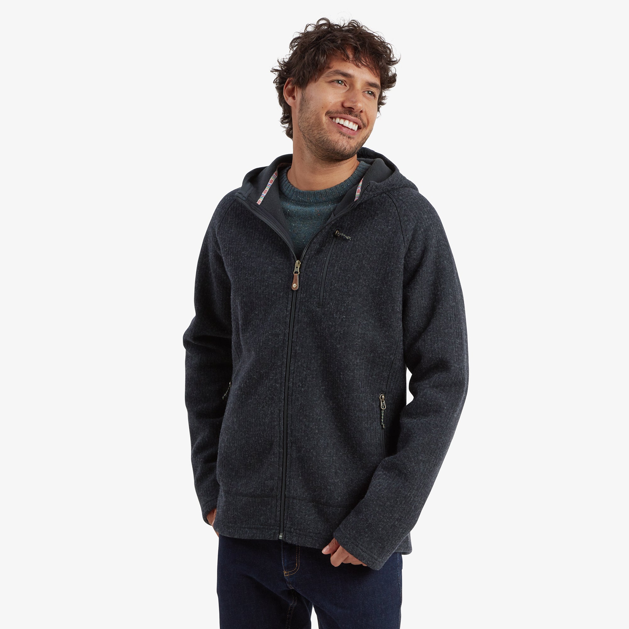 A smiling man models a Sherpa Adventure Gear Namgyal Hoodie in Black with a full-zip design. The jacket features a subtle heathered texture and a high neckline with a hood. He has his hands in the jacket's side pockets, paired with dark blue jeans and a casual stance.