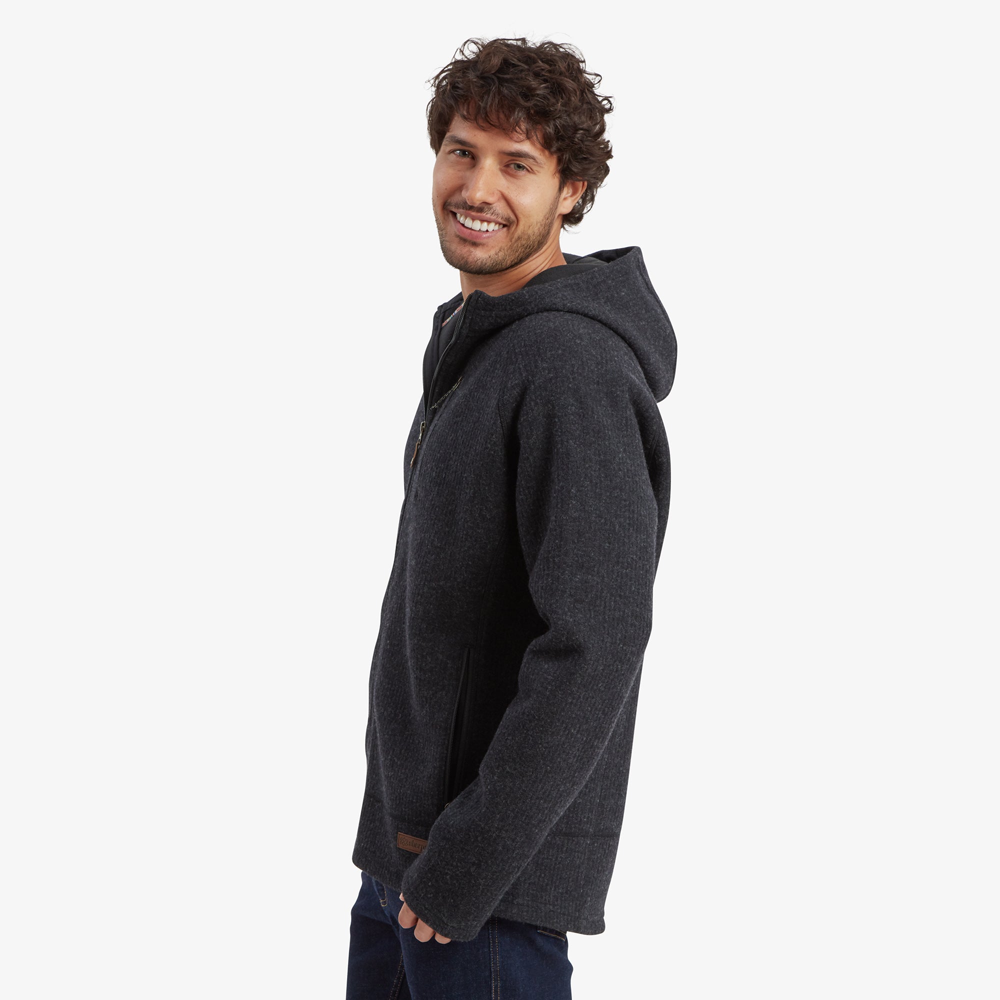 A side profile of the man wearing the Sherpa Adventure Gear Namgyal Hoodie in Black. The hood is shown resting on his back, and the sleeves and hem offer a tailored fit. He smiles toward the camera, highlighting the relaxed and functional design of the hoodie.