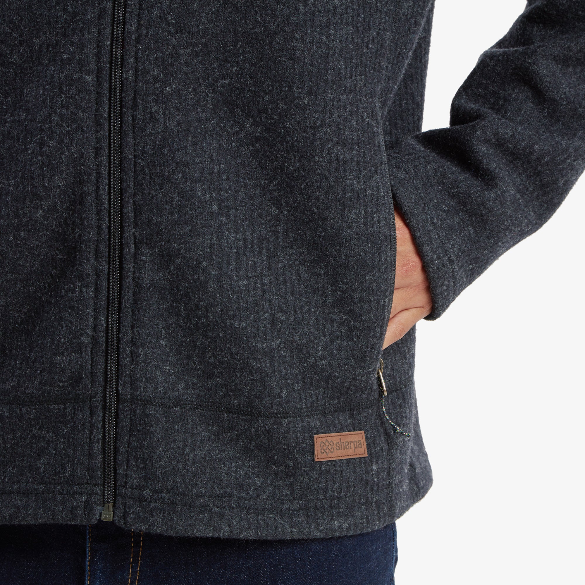 A zoomed-in look at the lower part of the Sherpa Adventure Gear Namgyal Hoodie in Black, featuring the Sherpa logo stitched on a leather patch near the hem. The image also displays the jacket's functional hand pockets and zippered closures, complemented by dark denim trousers.