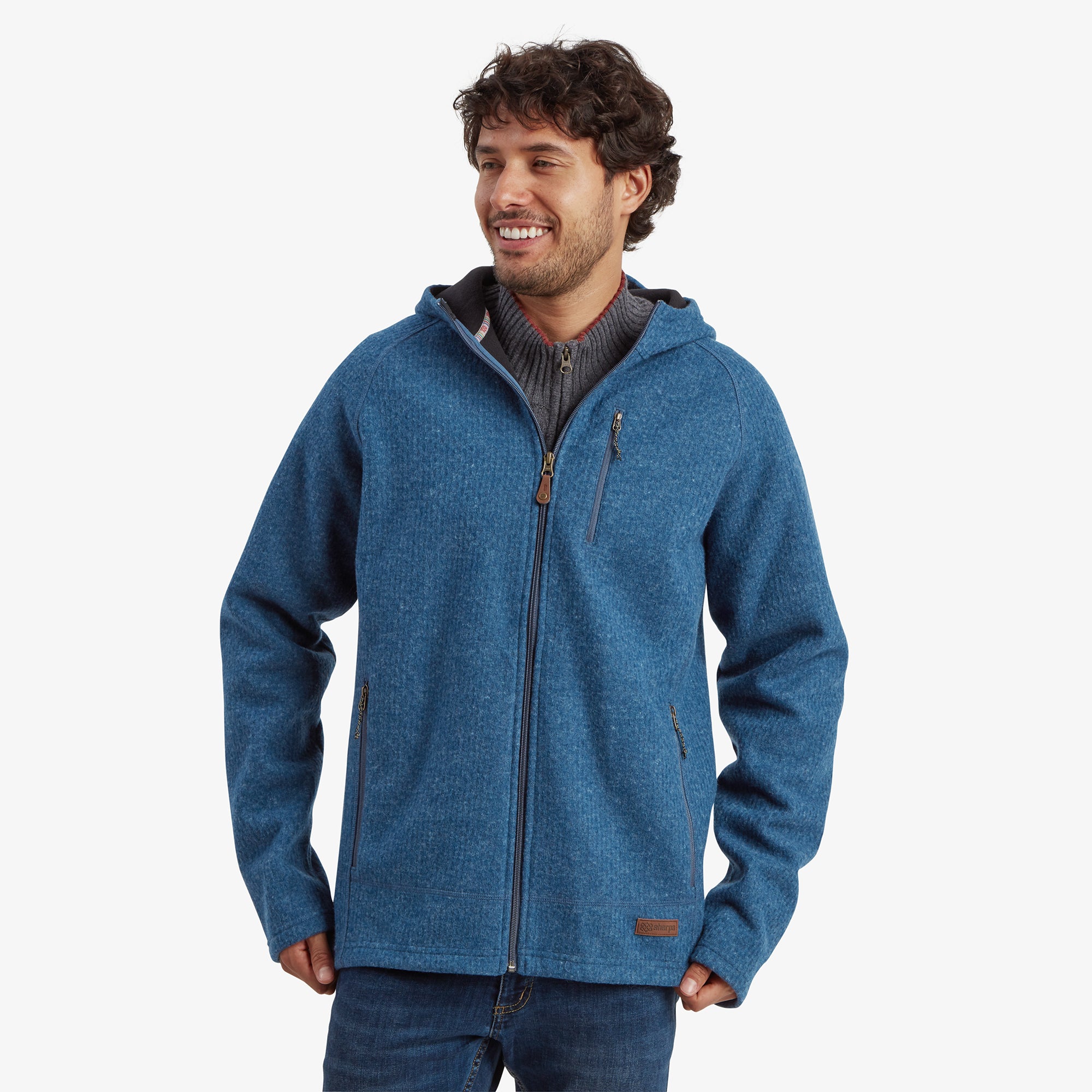 A smiling male model wearing a Sherpa Adventure Gear Namgyal Hoodie in Blue. The hoodie features a full zipper, a subtle knit texture, and a chest pocket with a vertical zipper. The model's hands are in the pockets, highlighting the practical design of the hoodie.