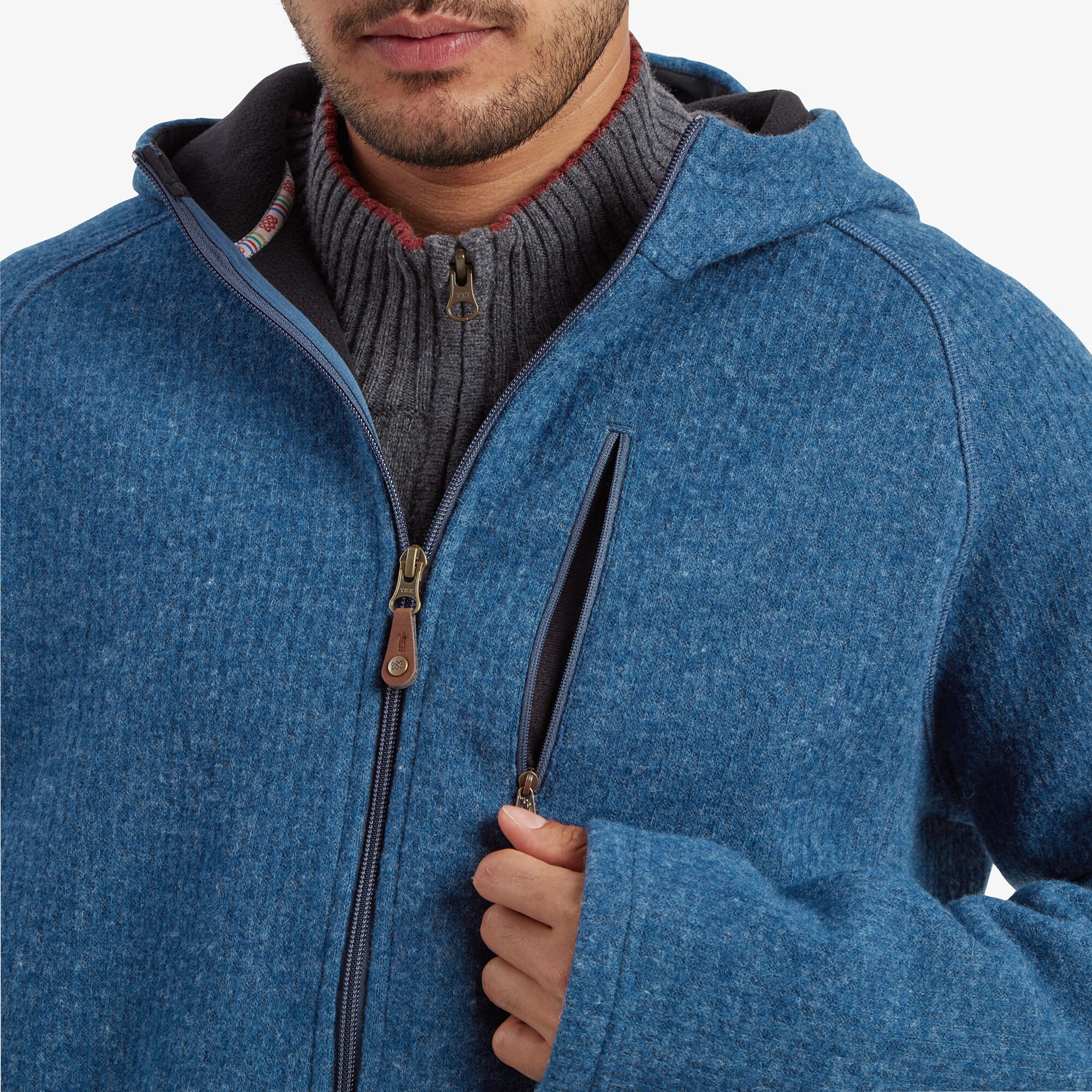 A close-up image of the model adjusting the Sherpa Adventure Gear Namgyal Hoodie in Blue’s chest pocket zipper. The texture of the blue knit fabric and the leather zipper pulls provide a sense of quality and craftsmanship.