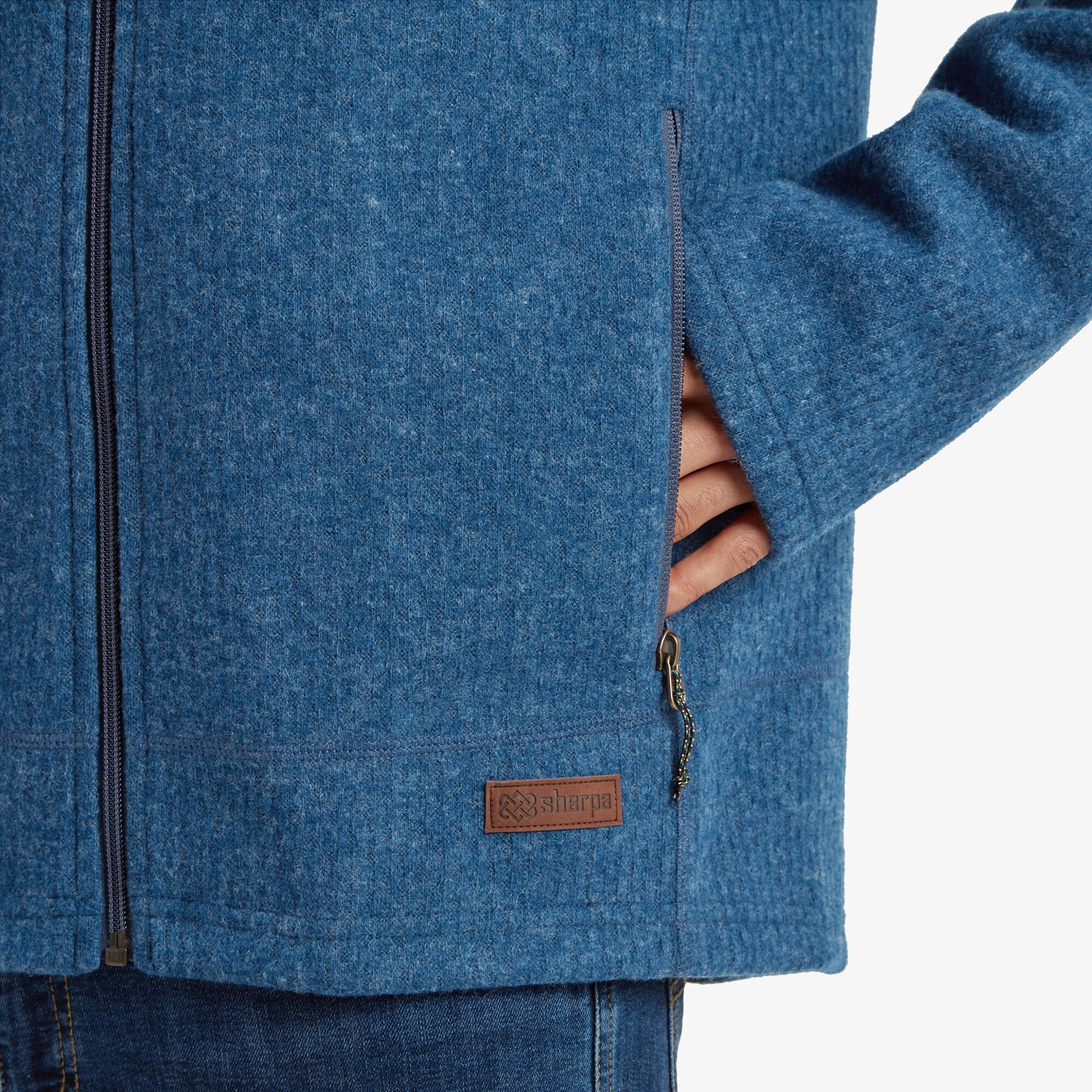 A close-up of the bottom left corner of the Sherpa Adventure Gear Namgyal Hoodie in Blue, showing the Sherpa logo on a leather patch. The side zipper pocket is partially open, emphasising the hoodie’s functionality and attention to detail.