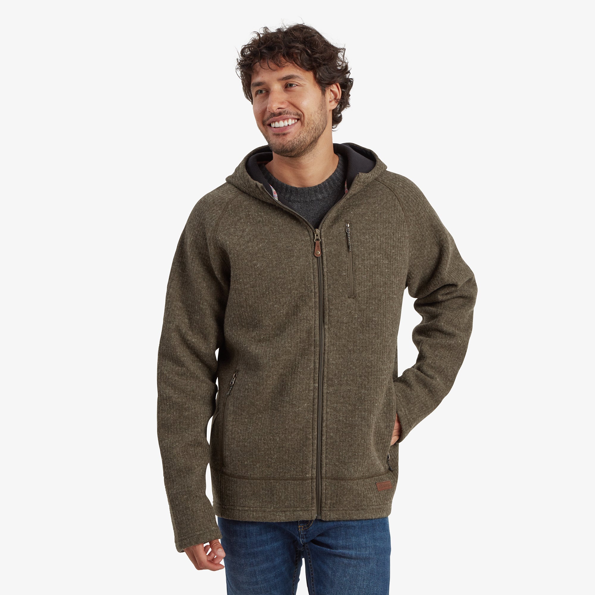 A man smiling while wearing a Sherpa Adventure Gear Namgyal Hoodie in Green. The hoodie features a full-length zipper, a small chest zip pocket, and a cozy fleece lining. The model has his hands in his pockets, showcasing the comfortable fit of the jacket.