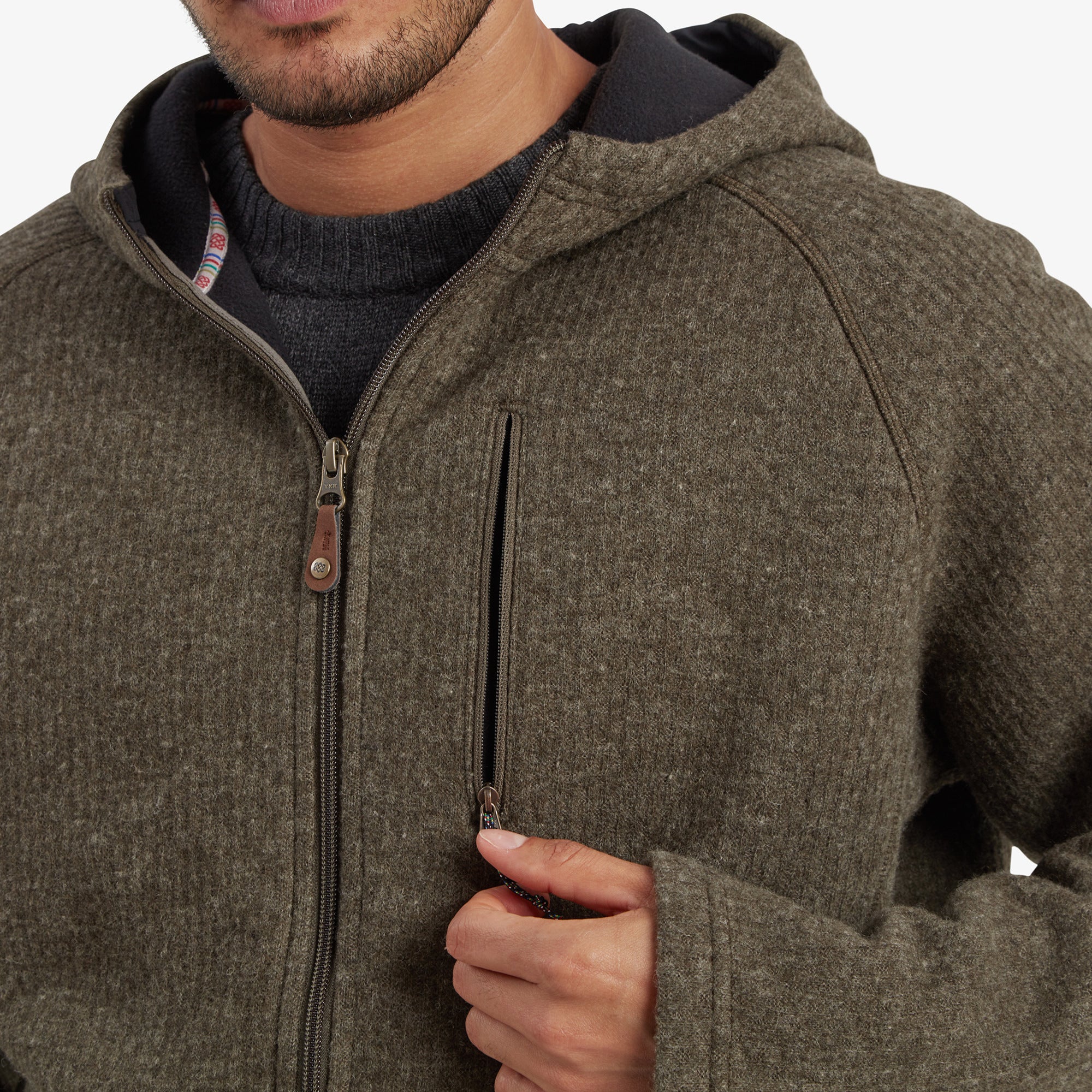 A close-up of the chest area of the Sherpa Adventure Gear Namgyal Hoodie in Green, focusing on the small zippered chest pocket, Sherpa branding, and the durable zipper pull. The texture of the fabric is highlighted in this detailed shot.