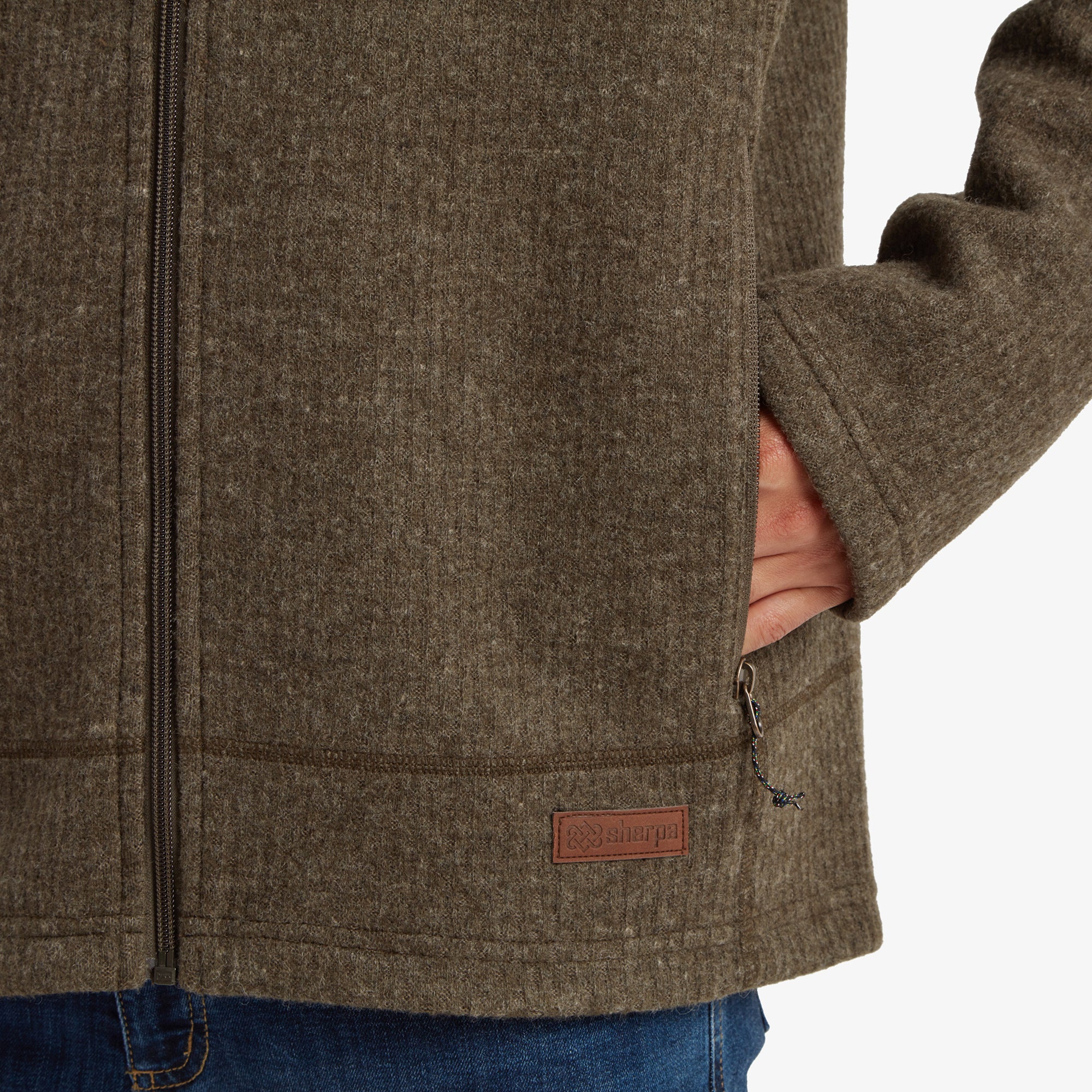 A close-up of the lower part of the Sherpa Adventure Gear Namgyal Hoodie in Green, focusing on the zippered hand pocket and the Sherpa leather logo patch stitched near the hem. The high-quality stitching and texture of the hoodie material are evident.