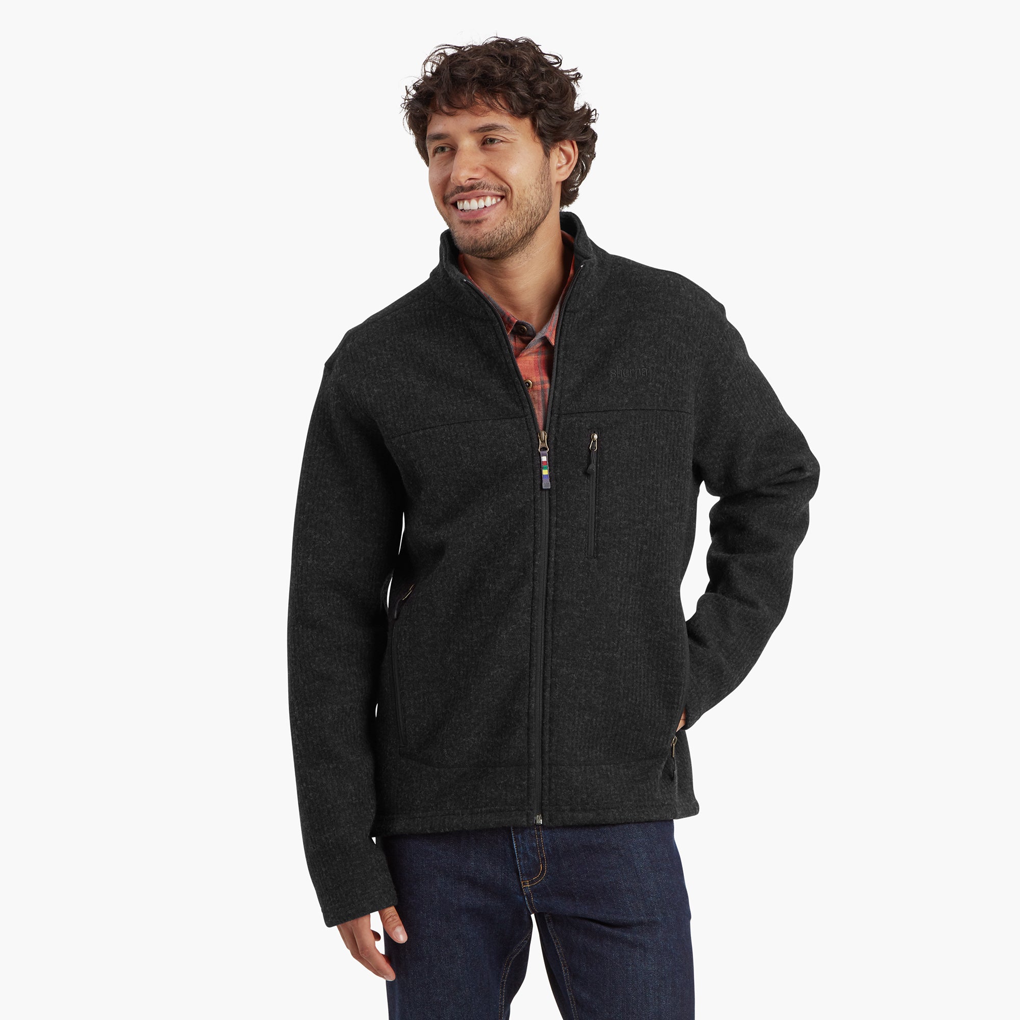 A man wearing a Sherpa Adventure Gear Namgyal Jacket in Black. The jacket features a full front zipper, a high collar, and a slim fit. The model is smiling and has one hand in the jacket pocket, showcasing its warmth and comfort.