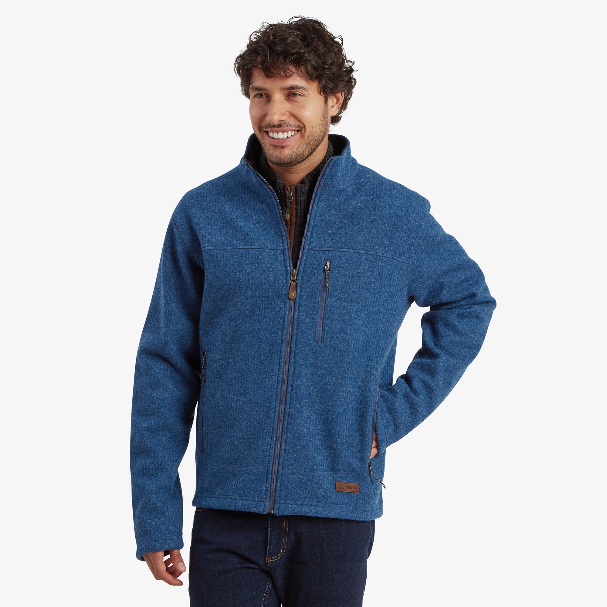 A man with medium-length curly brown hair and a light beard wears the Sherpa Adventure Gear Namgyal Jacket in Blue. The jacket is a full-zip style made from a textured knit fabric. It features a zippered chest pocket on the left side and two zippered hand pockets. The man is smiling with one hand resting in his pocket, and the background is a clean, off-white color.