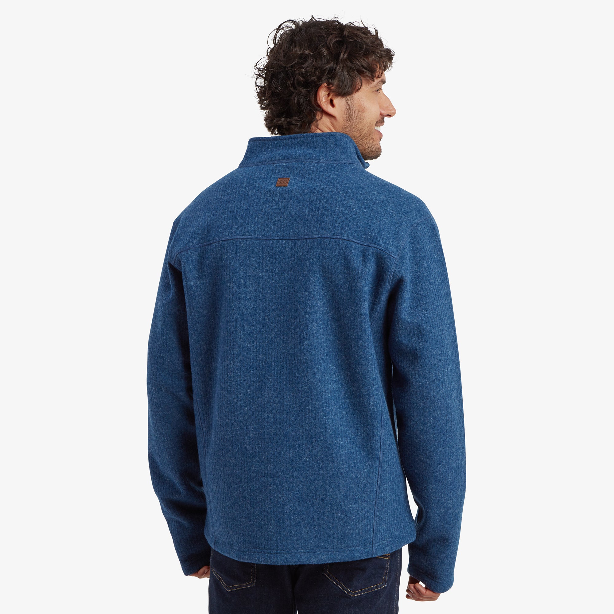 Back view of the Sherpa Adventure Gear Namgyal Jacket in Blue, showing a yoke seam across the upper back and a small Sherpa Adventure Gear logo patch at the center of the yoke. The textured knit fabric is consistent throughout. The model is slightly turned to the right with a smile, and the background is plain off-white.