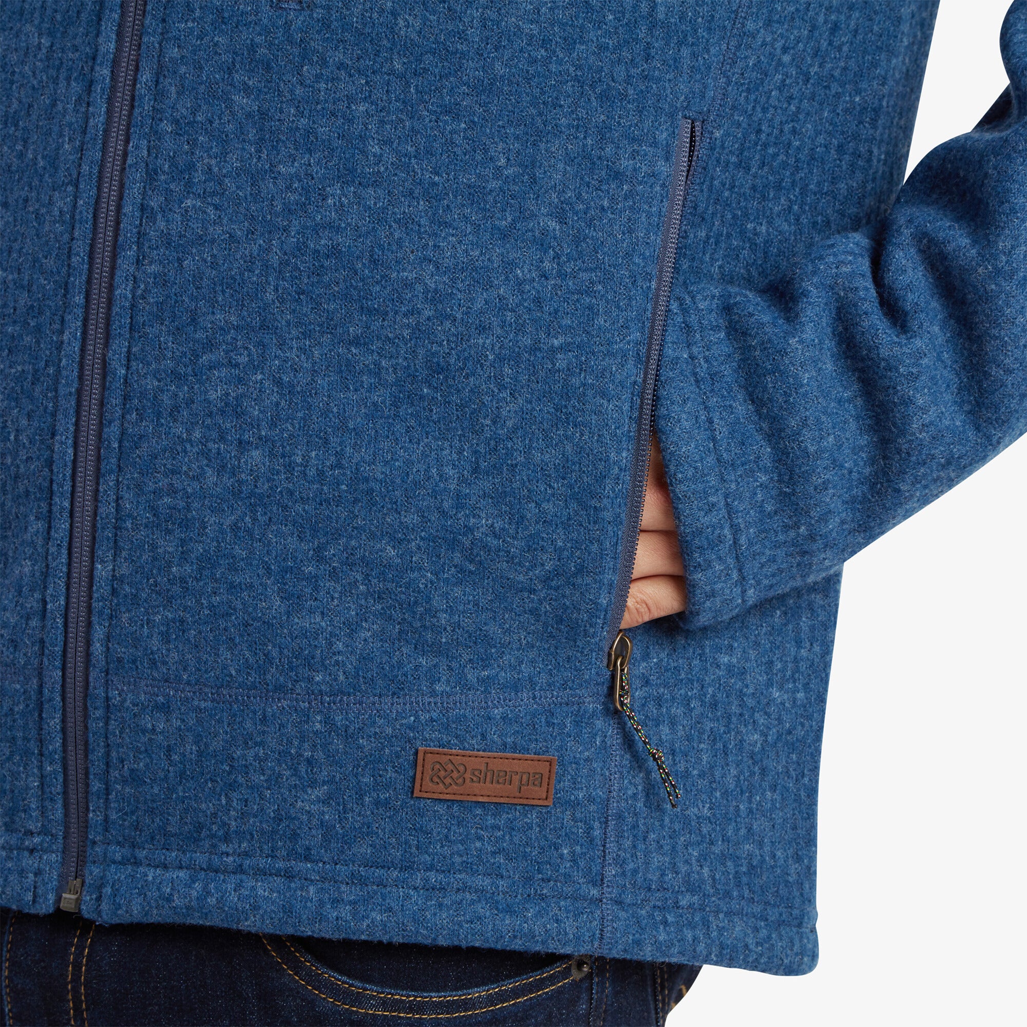 Close-up shot of the lower left side of the Sherpa Adventure Gear Namgyal Jacket in Blue, highlighting the texture of the knit fabric, the zippered hand pocket, and a brown leather Sherpa Adventure Gear logo patch stitched near the hem. The model’s hand is tucked into the pocket. The background is plain off-white.