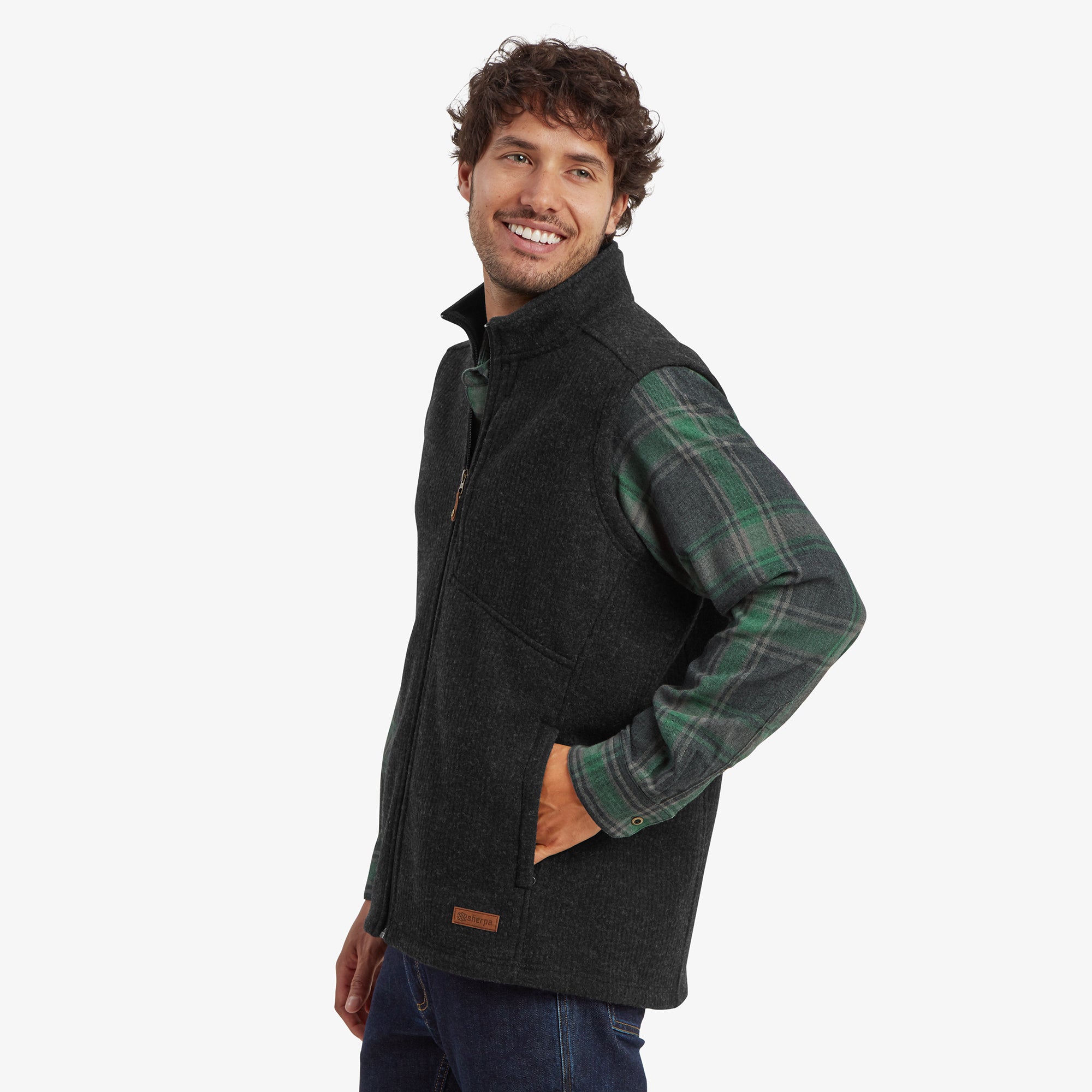 A side view of the Sherpa Adventure Gear Namgyal Vest in Black worn over the green plaid shirt. The model has one hand in the side pocket, highlighting the vest's practicality and snug fit. The outfit pairs well with dark blue jeans, giving an outdoorsy aesthetic.