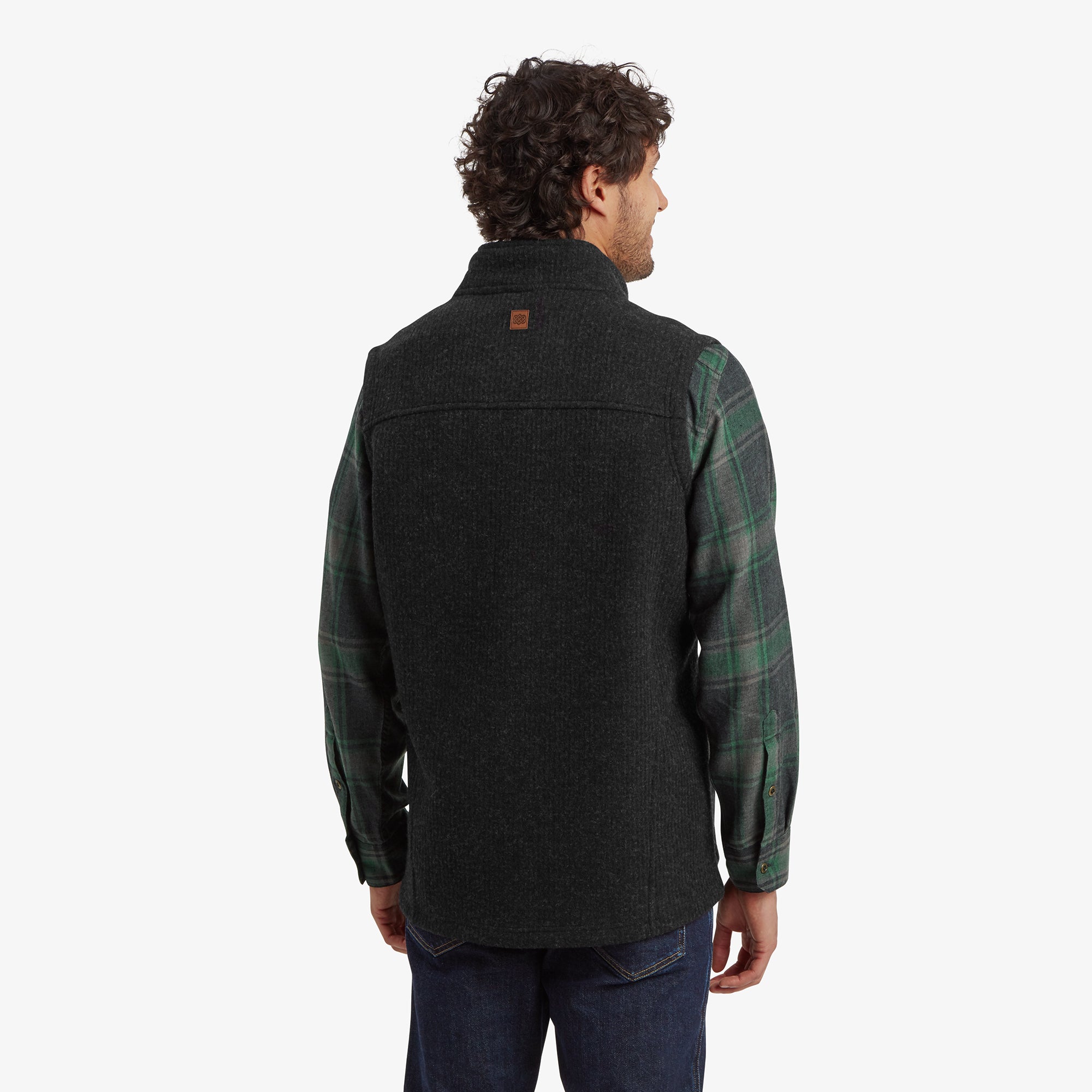 A back view of the Sherpa Adventure Gear Namgyal Vest in Black, showing its simple, streamlined design and subtle stitching. The high collar and the pairing with the green plaid shirt and jeans provide a clean, layered look suitable for cooler weather.