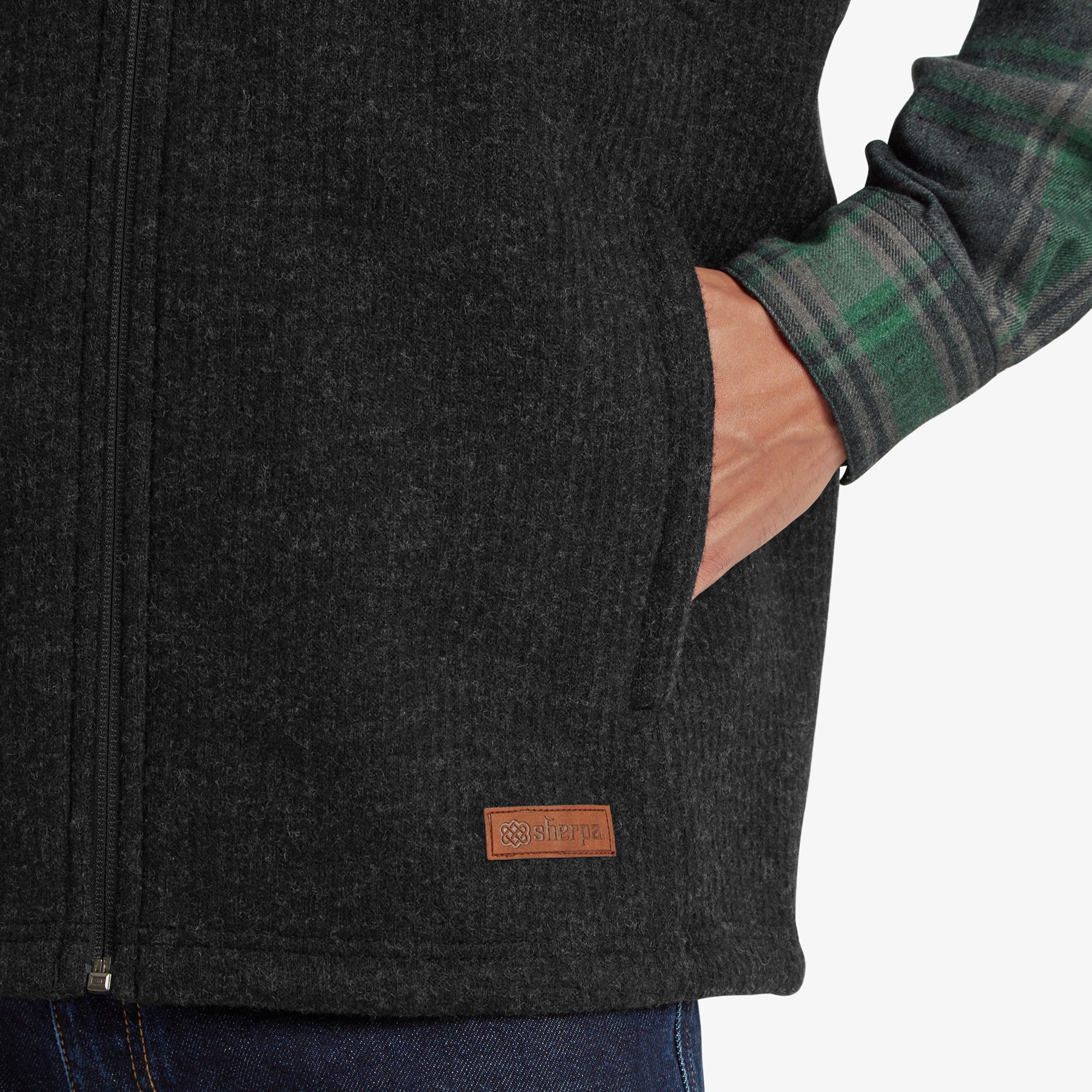 A close-up of the lower portion of the Sherpa Adventure Gear Namgyal Vest in Black. One hand is placed in the side pocket, and a small brown Sherpa Adventure Gear logo tag is visible near the hem. The pairing with dark blue jeans highlights the vest's ability to complement a casual wardrobe.