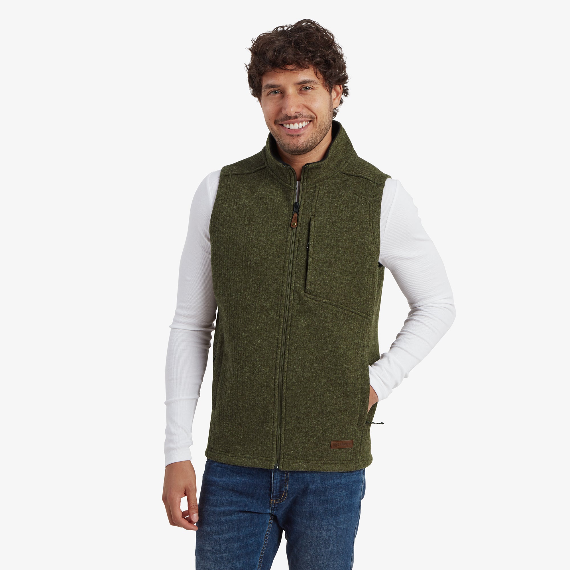 A male model wearing a Sherpa Adventure Gear Namgyal Vest in Green over a white long-sleeve shirt, paired with blue jeans. The vest features a high collar, a zippered chest pocket, and a relaxed fit. The model smiles warmly with his hands in the vest’s side pockets.