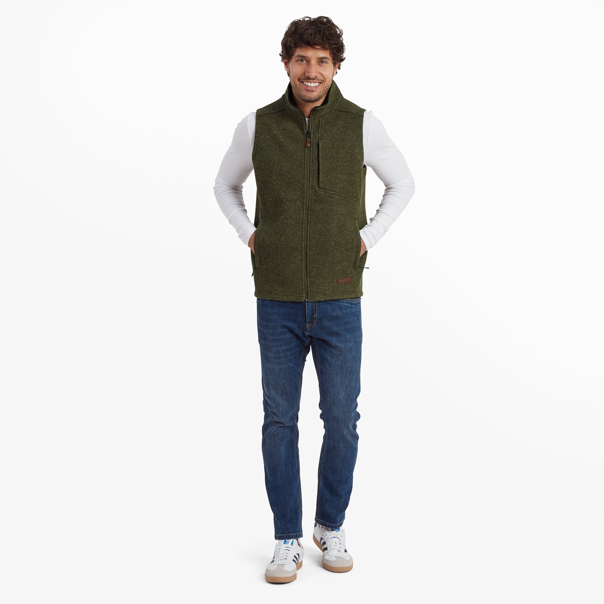 The same male model shown in a full-body pose. The Sherpa Adventure Gear Namgyal Vest in Green complements the white long-sleeve shirt and slim-fit blue jeans, paired with white casual trainers. The model exudes a casual and approachable style.