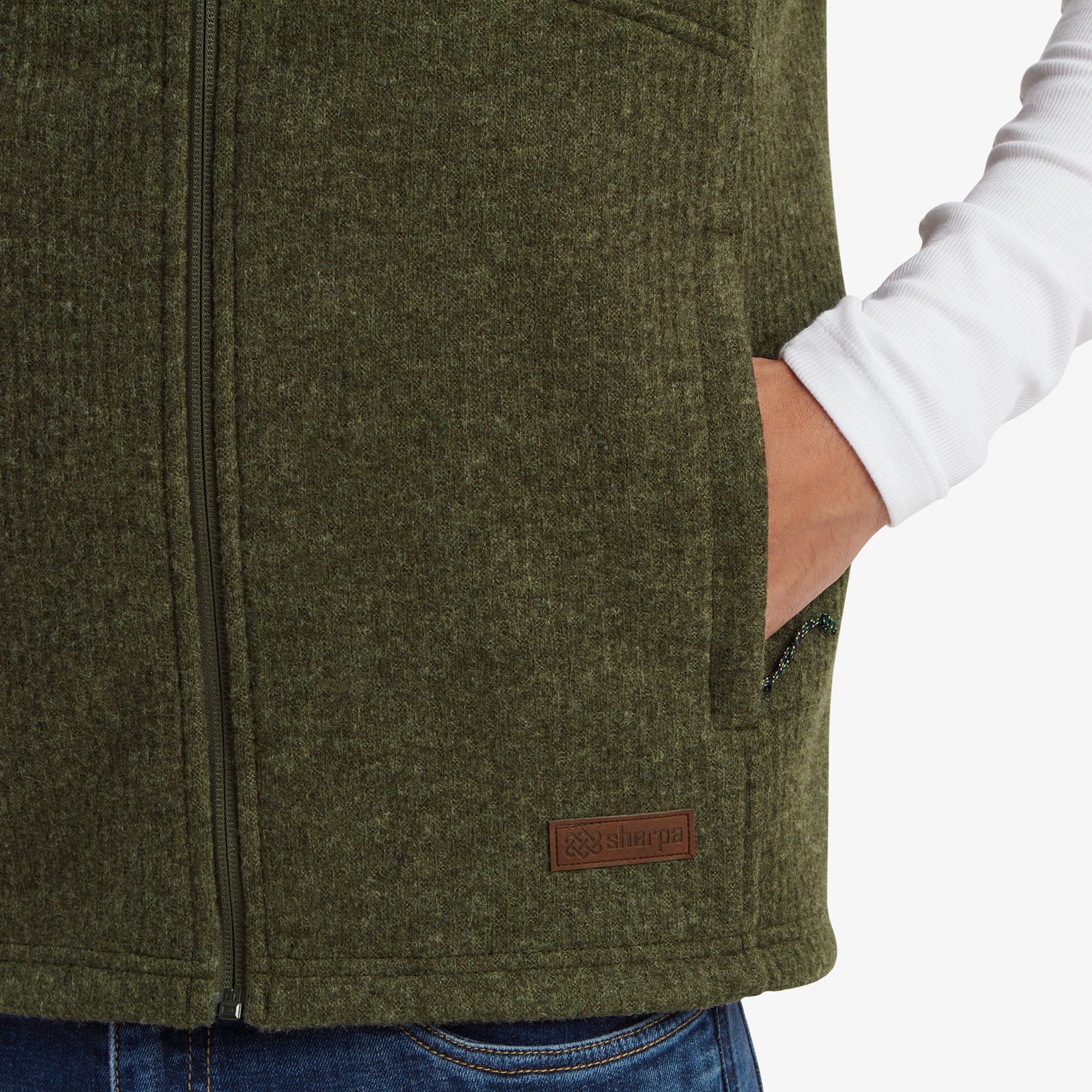 A close-up showing the Sherpa Adventure Gear Namgyal Vest in Green's side pocket and the Sherpa logo patch stitched near the hem. The texture and craftsmanship of the fabric are emphasised, giving a sense of premium quality. The model’s hand rests casually in the pocket.