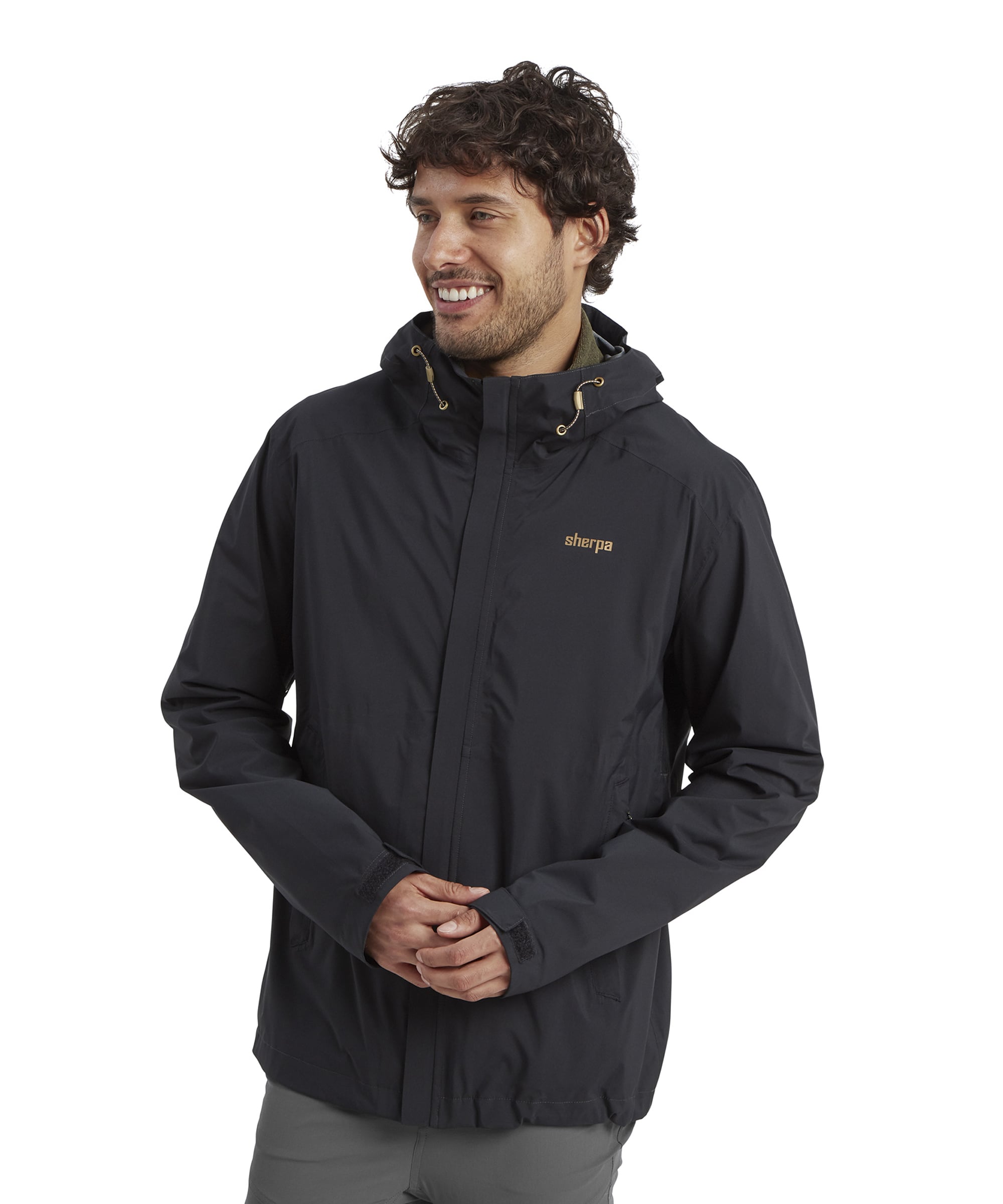 A man wearing a Sherpa Adventure Gear Nima 2.5-Layer Jacket in Black with a subtle sheen. The jacket features a hood with drawstrings, Sherpa's logo on the chest in gold, and a zippered front closure. The model smiles and clasps his hands in front.