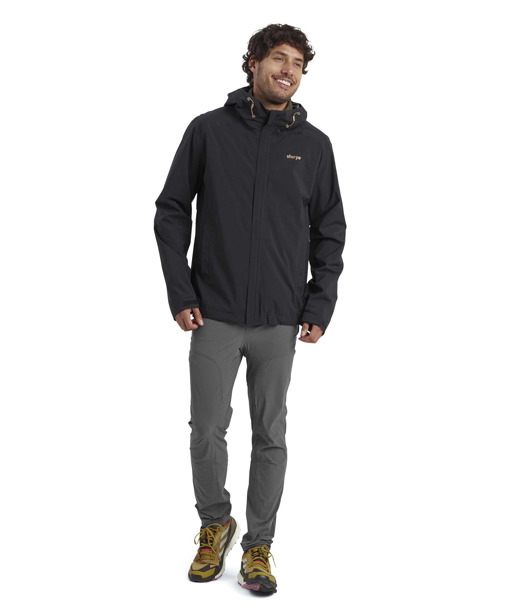 A full-body view of the model showcasing the Sherpa Adventure Gear Nima 2.5-Layer Jacket in Black paired with gray outdoor trousers and yellow hiking shoes. The man stands confidently, giving a casual pose with a relaxed smile.