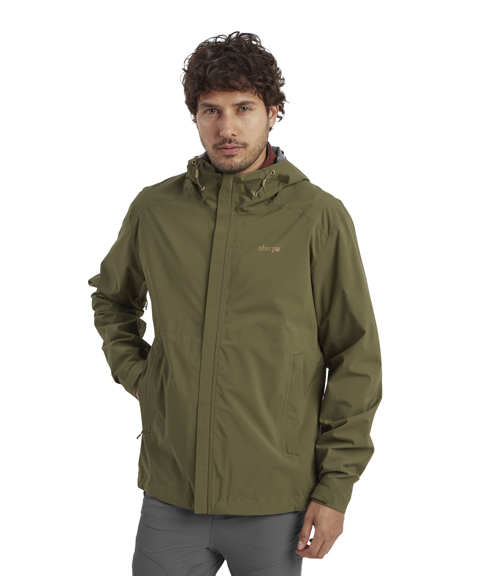 A front-facing view of a man wearing the Sherpa Adventure Gear Nima 2.5-Layer Jacket in Green. The jacket features a sleek, minimalist design with a high collar and a subtle Sherpa logo on the left chest. The man has his hands casually tucked into the jacket’s front pockets, highlighting its practicality.