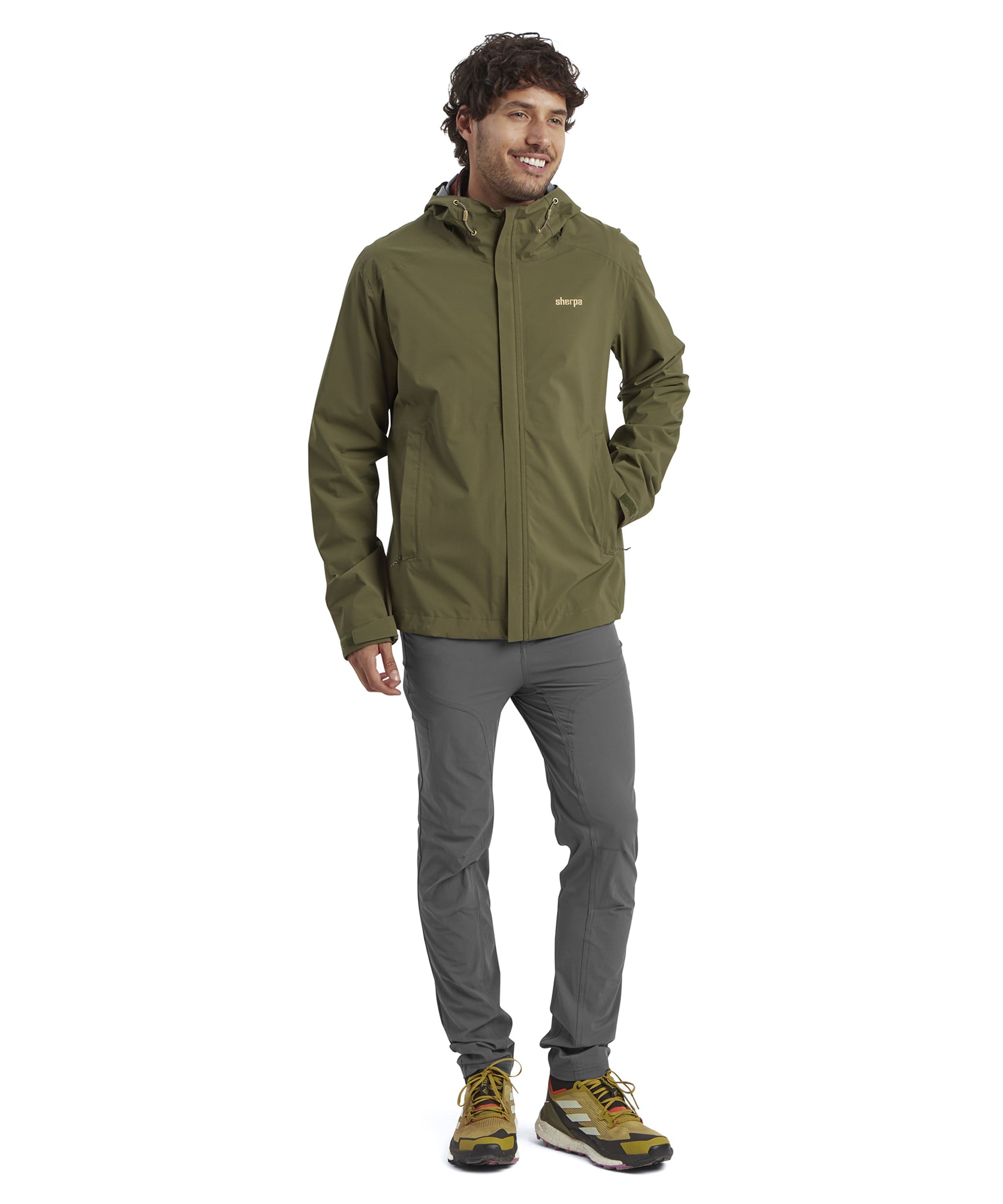 A full-body shot of the man modeling the Sherpa Adventure Gear Nima 2.5-Layer Jacket in Green, paired with gray trousers and yellow hiking shoes. He stands casually with a cheerful smile, showcasing the jacket’s comfortable fit and outdoor-ready look.