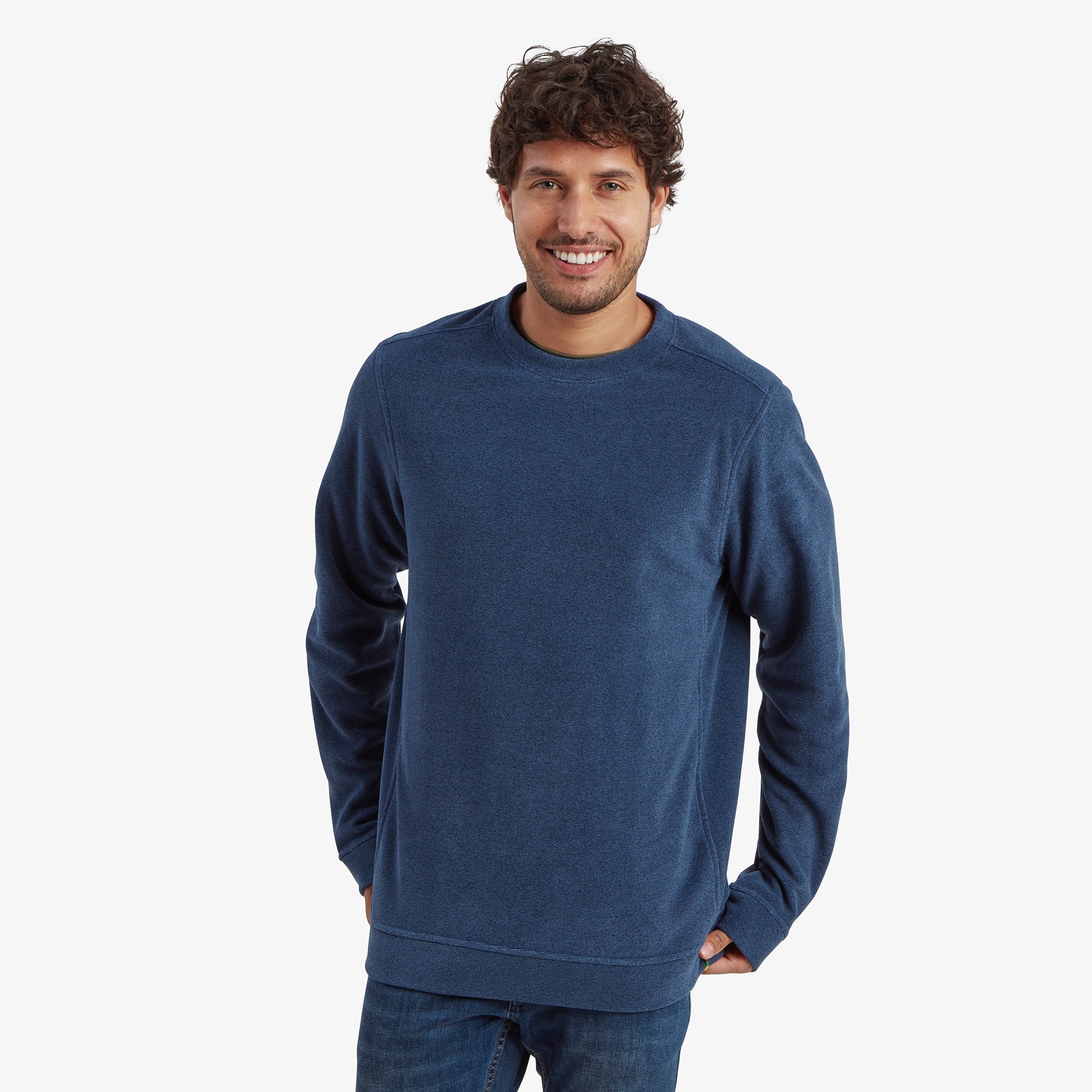 A man wearing a Sherpa Adventure Gear Rolpa Eco Crew in Blue, standing against a plain white background. The sweatshirt has a relaxed fit, featuring a clean and minimal design with subtle shoulder seams. The model is smiling warmly, showcasing the comfortable and casual nature of the garment.