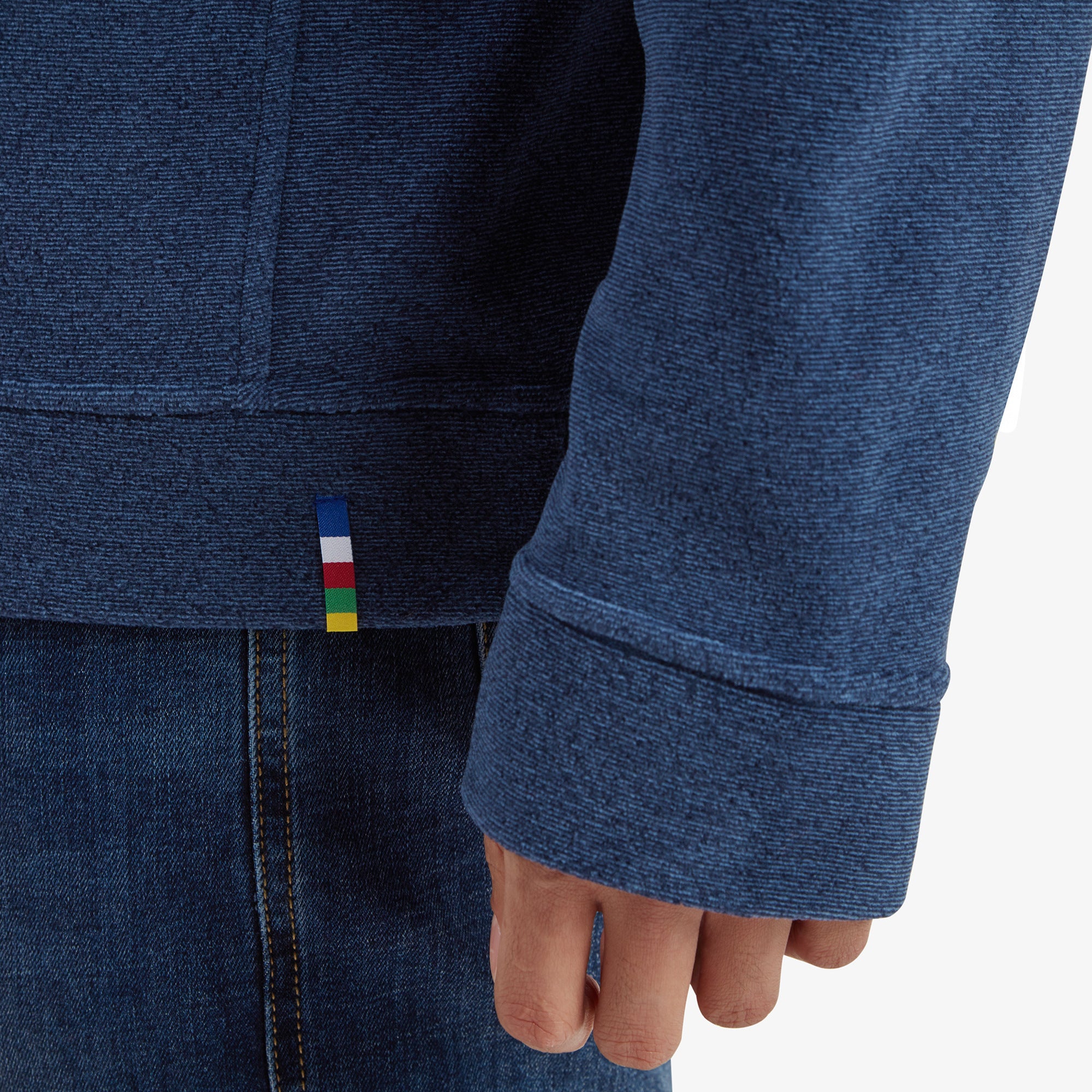 A close-up of the hem and sleeve cuff of the Sherpa Adventure Gear Rolpa Eco Crew in Blue, showing the fine textured knit of the fabric. A colorful woven detail in the form of a small tag, featuring stripes of blue, red, yellow, and green, adds a playful accent to the minimalist design. The shot emphasises the attention to detail and quality craftsmanship of the garment.