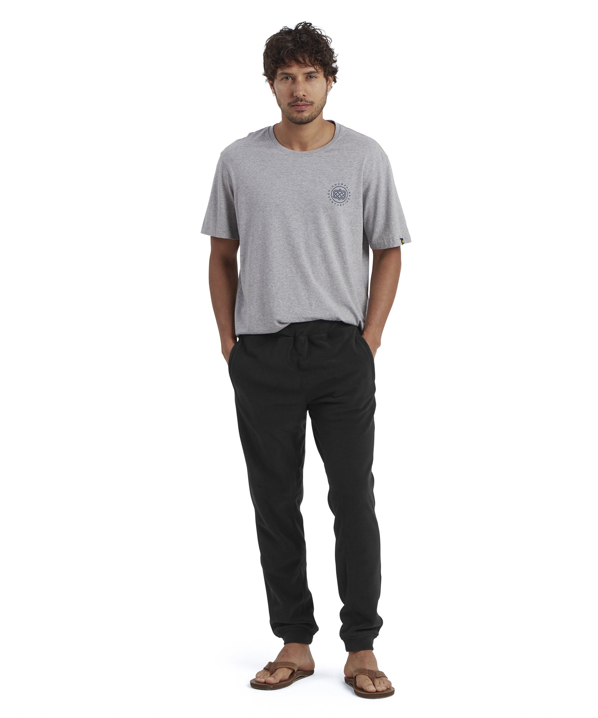 A full-body image of a man wearing a gray T-shirt paired with Sherpa Adventure Gear Rolpa Eco Fleece Jogger in Black. The trousers are styled with a casual fit and elasticated cuffs, complemented by brown flip-flops. The model stands with a relaxed posture, hands in pockets, showcasing a laid-back outfit.