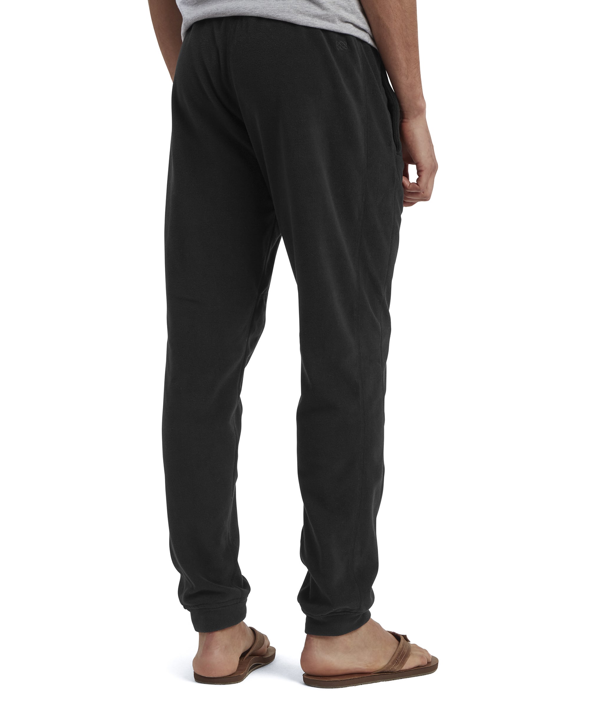 A back view of the Sherpa Adventure Gear Rolpa Eco Fleece Jogger in Black, capturing the elastic waistband and the overall casual fit. The model's flip-flops and light gray T-shirt add to the minimalistic, comfortable styling of the trousers.