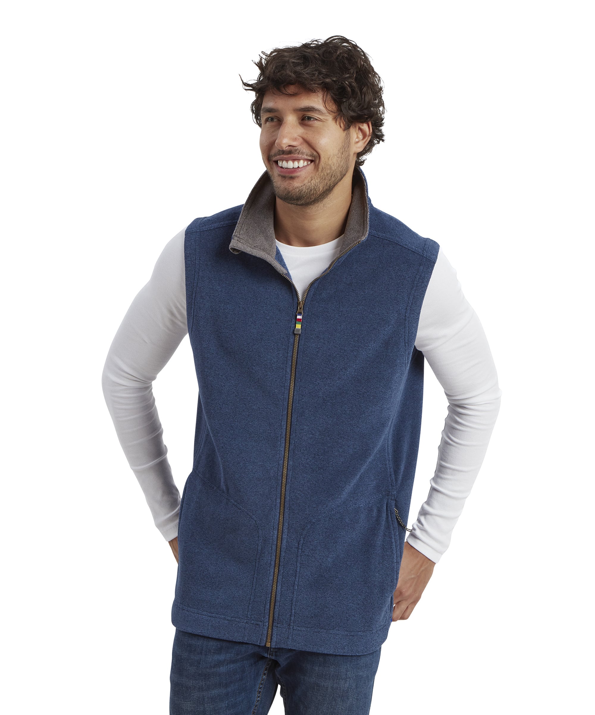 A man with curly brown hair and a beard wears a Sherpa Adventure Gear Rolpa Eco Vest in Blue over a white long-sleeved shirt. He is smiling with his hands in the pockets of the vest, looking off to the side. The vest features a front zipper and a raised collar with a soft grey lining.