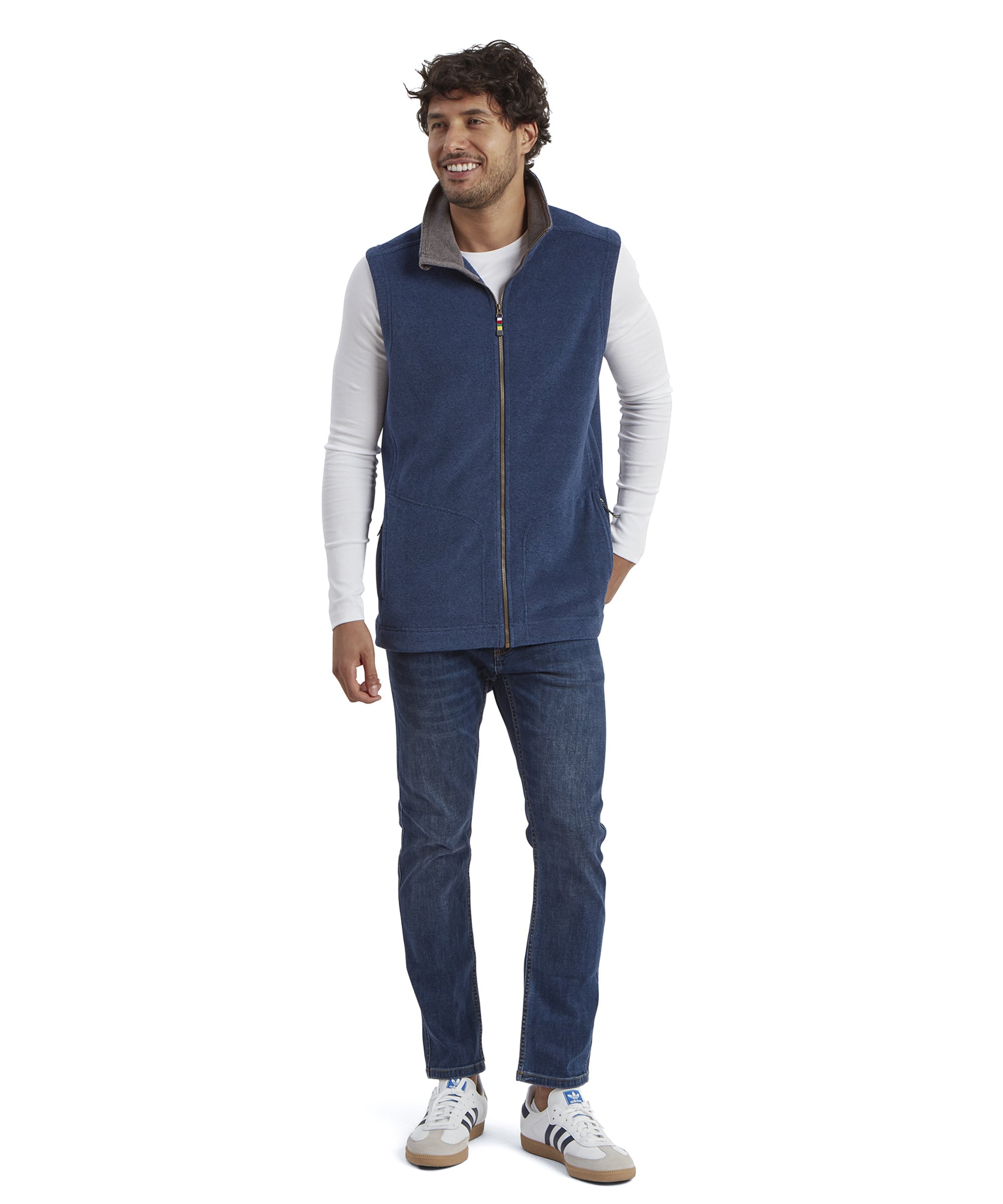 The same man stands in a relaxed pose wearing the Sherpa Adventure Gear Rolpa Eco Vest in Blue paired with dark jeans and white trainers. The full-body shot highlights the vest’s comfortable fit and casual styling, with the zipper fully closed and the collar raised.