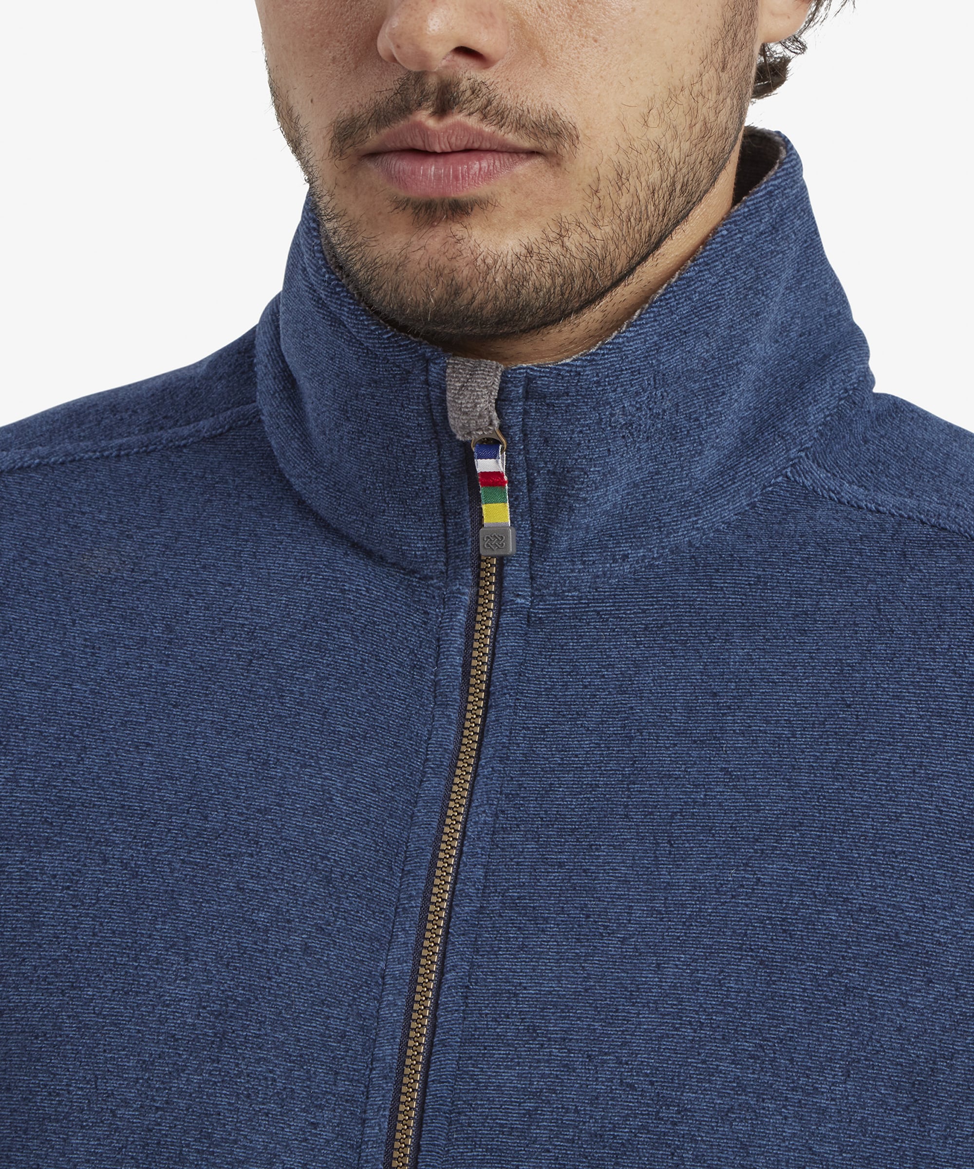 A close-up shot of the upper part of the Sherpa Adventure Gear Rolpa Eco Vest in Blue, highlighting the front zipper, which has a decorative fabric pull tab in multicolored stripes. The raised collar is neatly structured, with a grey inner lining contrasting the blue fleece exterior.