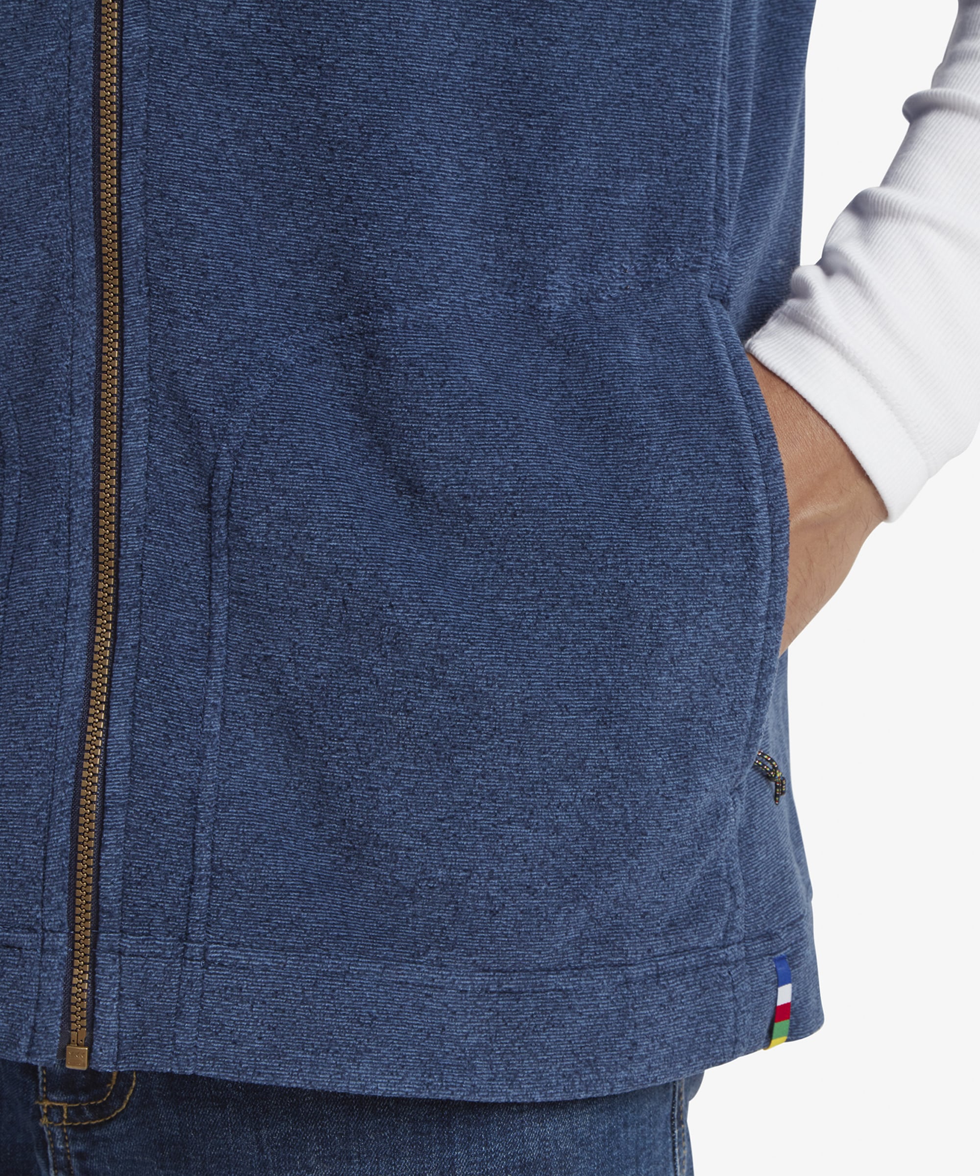 A detailed close-up of the lower front portion of the Sherpa Adventure Gear Rolpa Eco Vest in Blue, focusing on the hand pockets. The zipper and stitching details are sharp and clean, and a small logo tag is sewn near the pocket opening. The man’s hand is resting inside the pocket, showcasing its practical design.