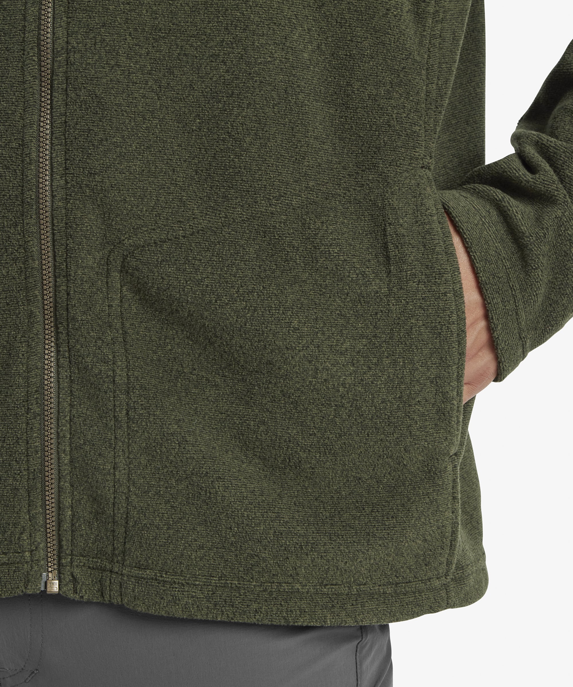 A close-up of the lower section of the Sherpa Adventure Gear Rolpa Eco Jacket in Green, highlighting the front pocket with a hand casually resting inside. The texture of the fleece material and the structured hemline are prominently displayed.