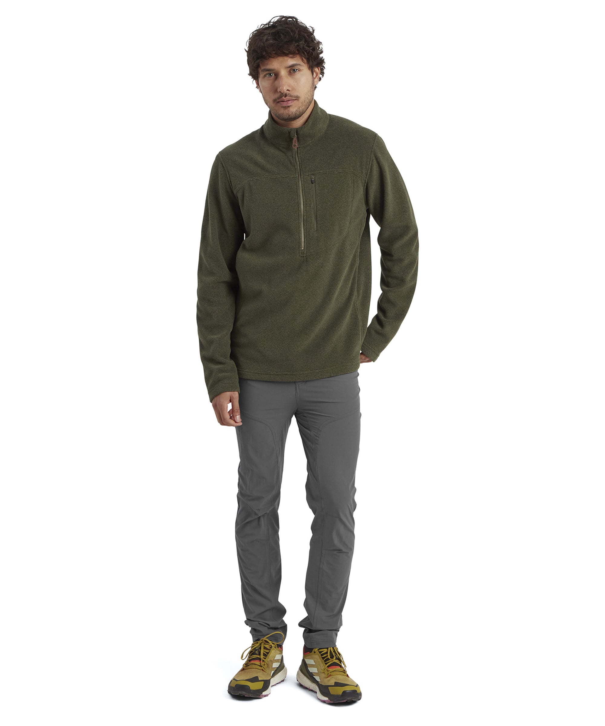 A full-body shot of the same man modeling the Sherpa Adventure Gear Rolpa Eco Quarter Zip in Green. He pairs the pullover with slim-fit gray trousers and yellow trail shoes, striking a relaxed pose with one hand in his pocket. The outfit presents a sporty and functional look, suitable for hiking or casual outings.