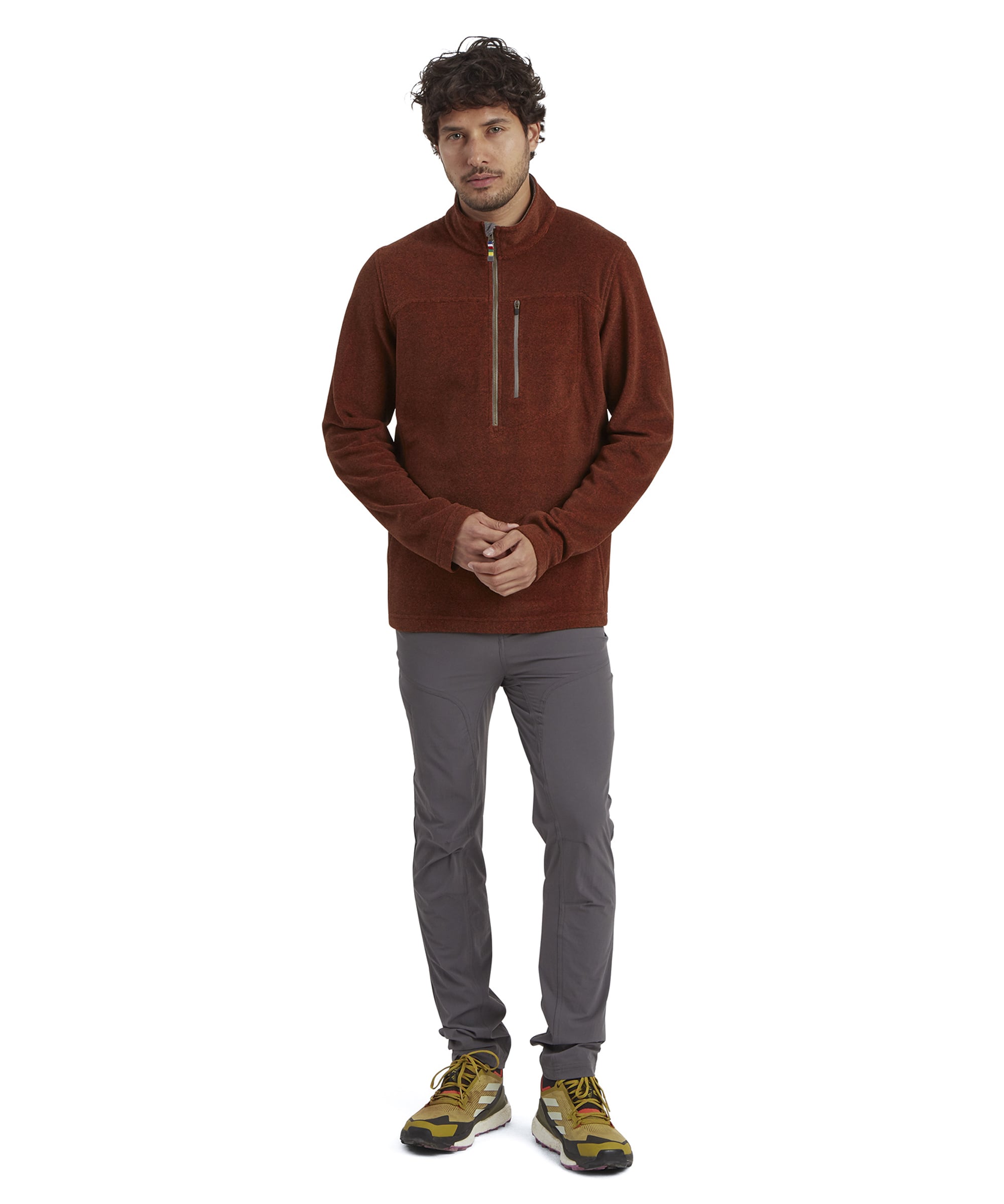 A full-body view of the man wearing the Sherpa Adventure Gear Rolpa Eco Quarter Zip in Red paired with gray pants and trail-inspired shoes with yellow accents. He is standing upright with his hands clasped loosely, presenting a casual and outdoor-ready style.