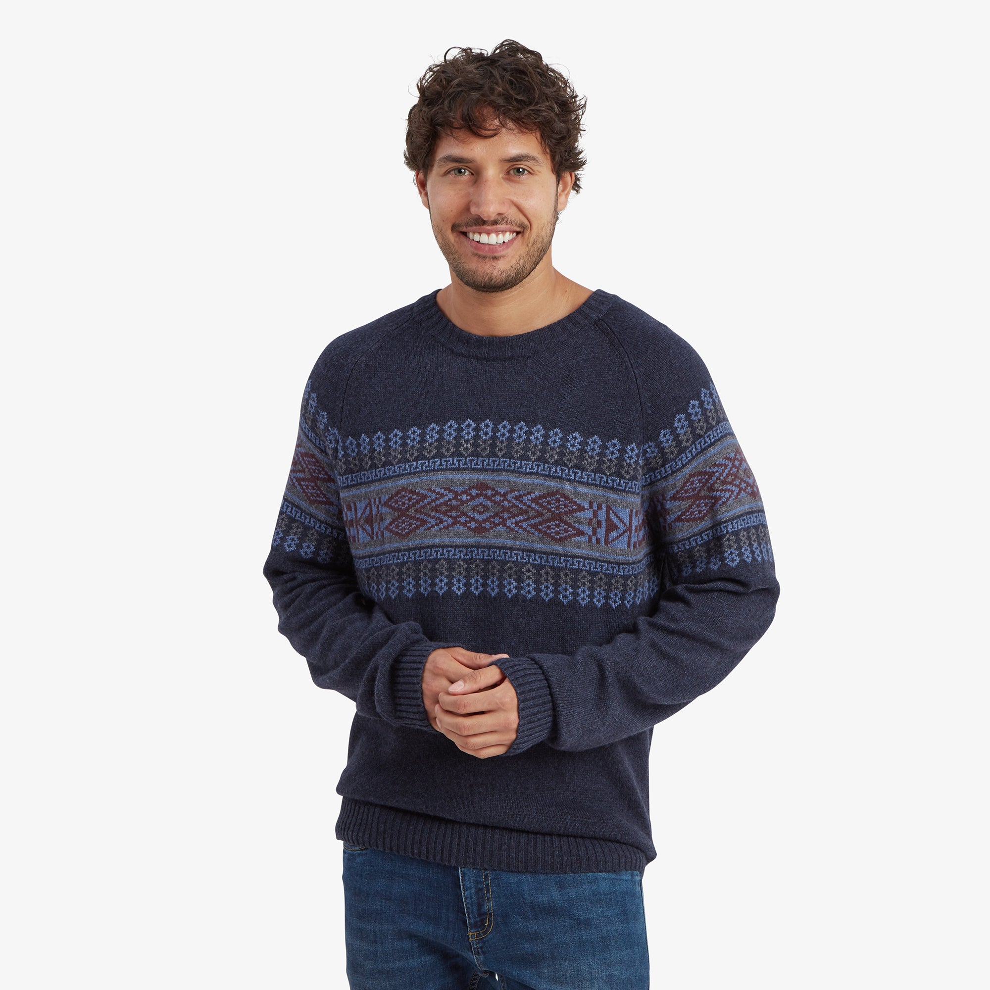 A man wearing a Sherpa Adventure Gear Sajit Eco Crew in Blue with intricate geometric patterns in shades of blue and burgundy. He is smiling and has his hands clasped in front of him. The sweater has ribbed cuffs and a hem. He is paired with blue denim jeans.