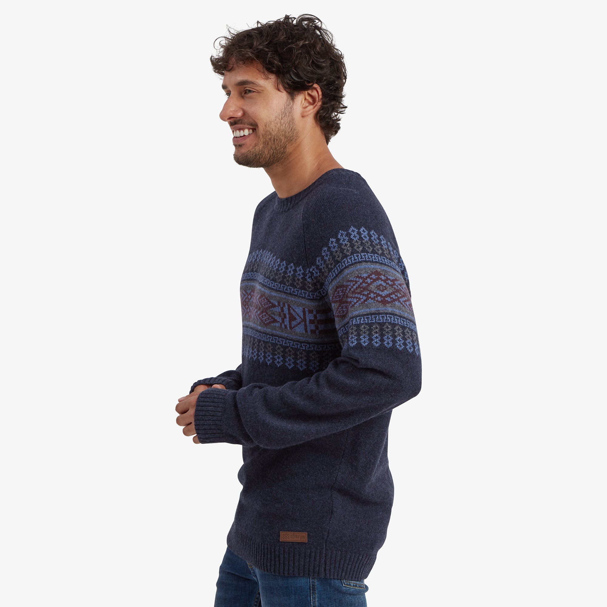 The man is turned slightly to the left, showcasing the detailed side profile of the Sherpa Adventure Gear Sajit Eco Crew in Blue's geometric pattern. His left hand is casually hanging by his side, and he is smiling.