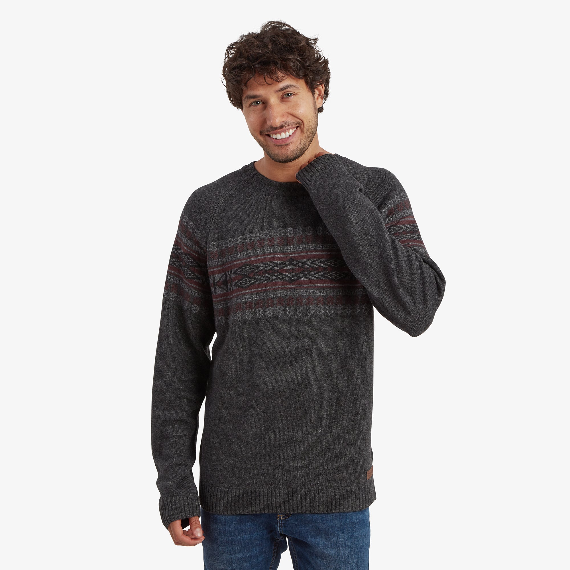 A smiling man wearing a Sherpa Adventure Gear Sajit Eco Crew in Grey with intricate horizontal patterns in shades of red and grey across the chest and arms. He stands casually with one hand near his face, showcasing the sweater's cozy texture and relaxed fit.