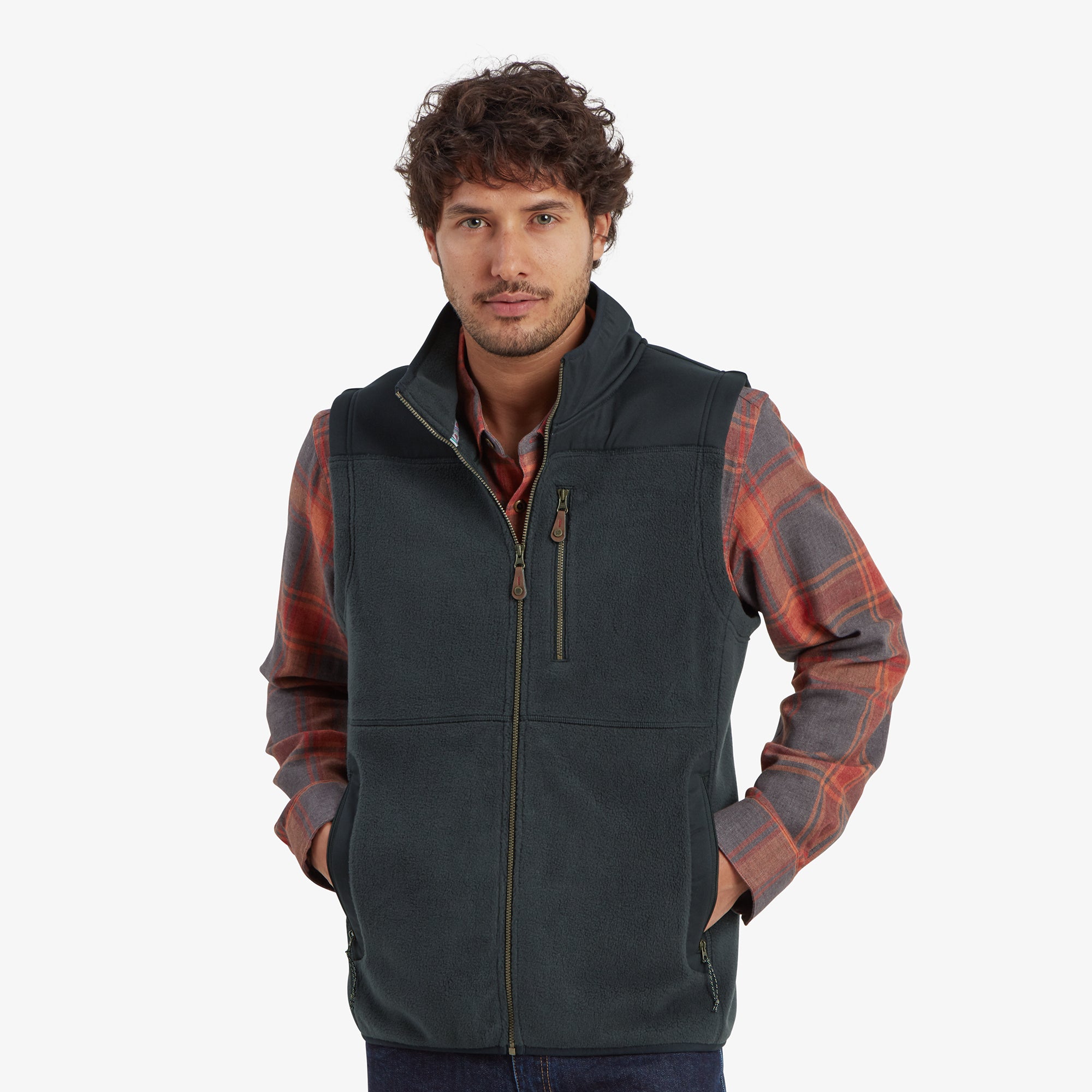 A man is shown wearing a Sherpa Adventure Gear Sanani Eco Fleece Vest in Black over a plaid shirt. The vest features a high collar, a zippered chest pocket, and two zippered hand pockets. He has his hands casually placed in the vest pockets, standing in a neutral pose.