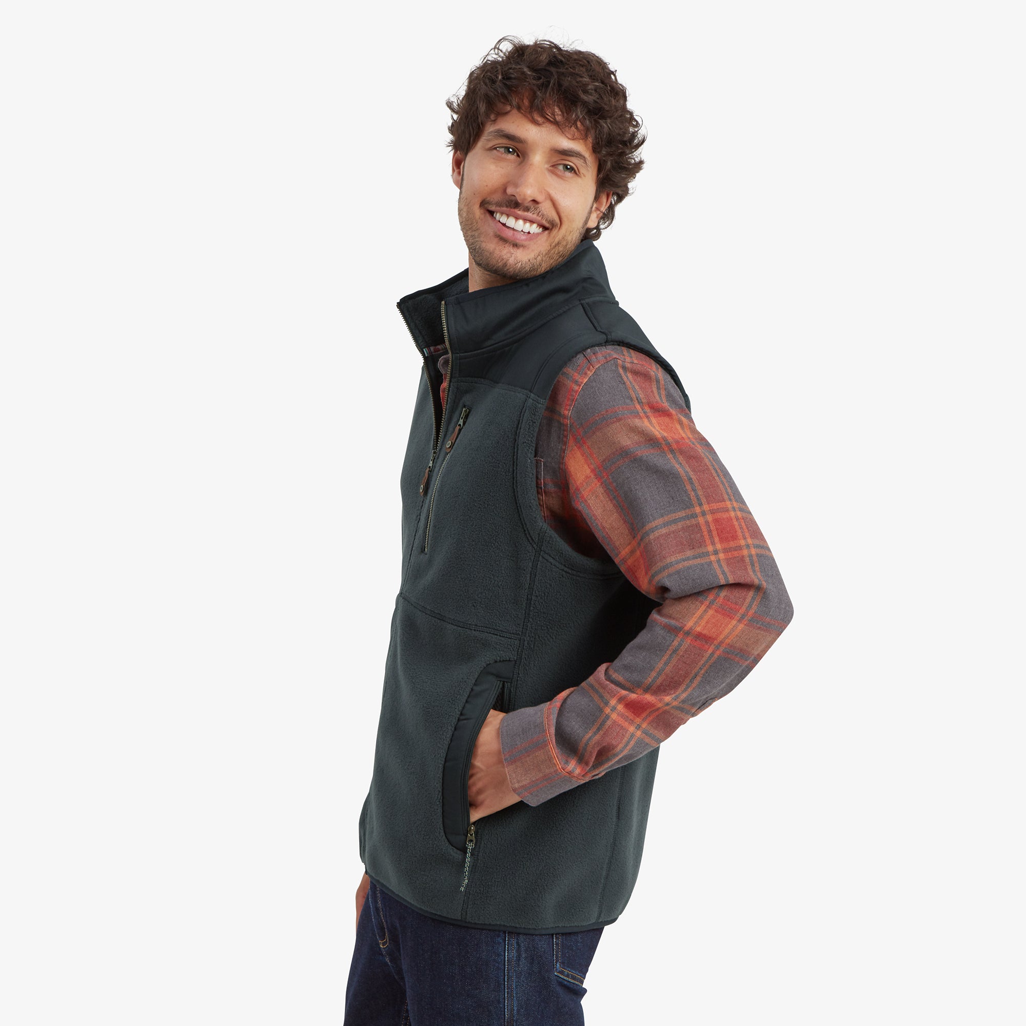 A side profile of the man wearing the Sherpa Adventure Gear Sanani Eco Fleece Vest in Black. The zippered hand pockets and the fit of the vest along the side are clearly visible. He looks cheerful, slightly turning his head toward the camera.