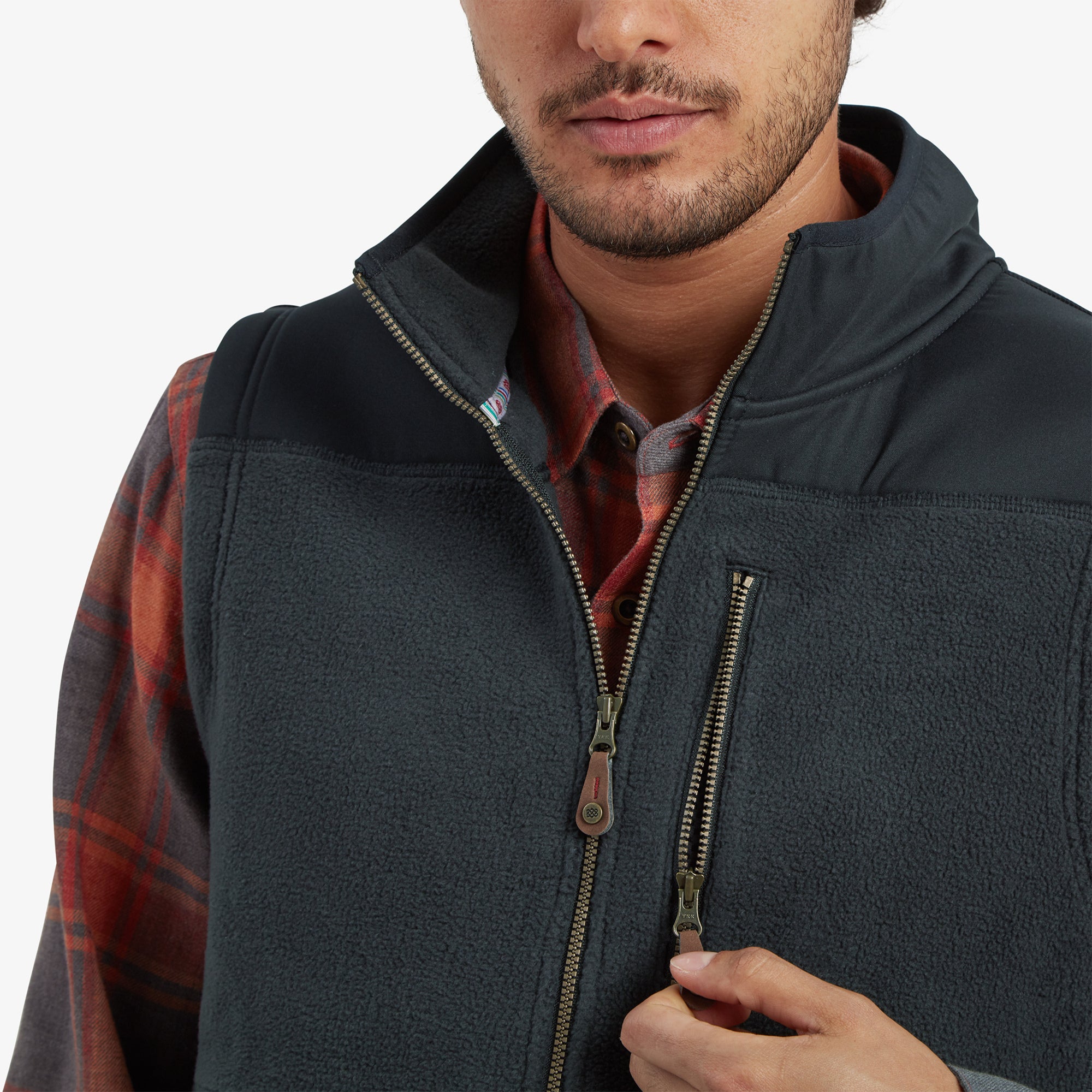 A close-up of the zippered chest pocket and high collar of the Sherpa Adventure Gear Sanani Eco Fleece Vest in Black. The man’s hand is shown holding the zipper pull, demonstrating the utility of the pocket. The plaid pattern of the shirt underneath is also partially visible.