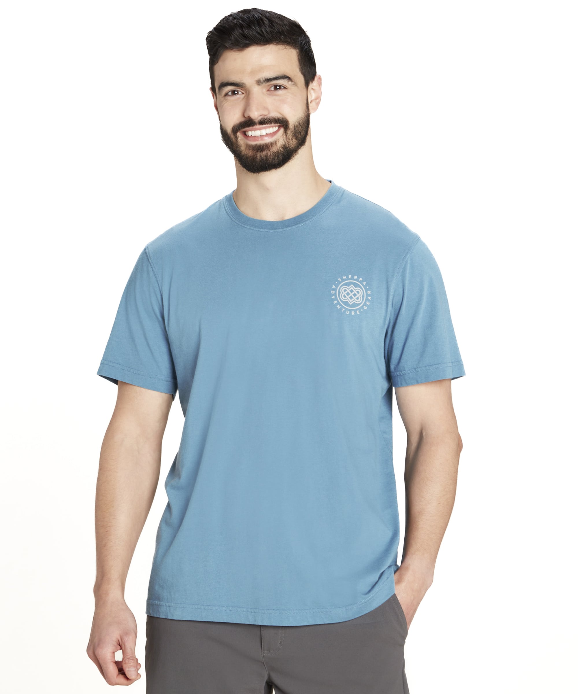 A man wearing a Sherpa Adventure Gear Summit T-Shirt in Blue with short sleeves, featuring a small Sherpa Adventure Gear logo in white on the left chest area. He is smiling, standing in a relaxed pose, and paired with gray shorts in front of a neutral background.